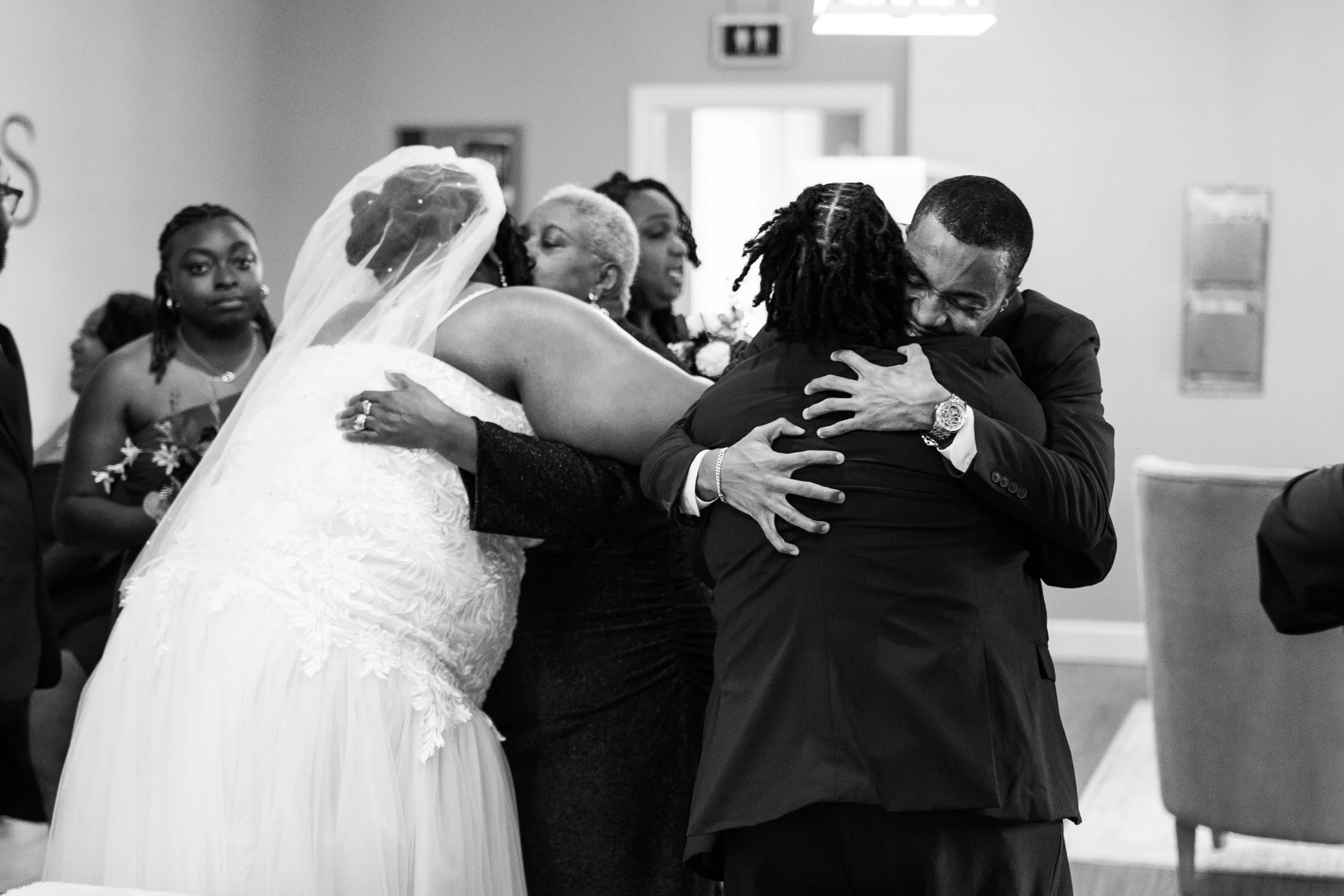 A group of people at a wedding celebration, with a woman in a white wedding dress and veil hugging a man in a black suit, surrounded by others dressed formally at Uwharrie Vineyards wedding venue