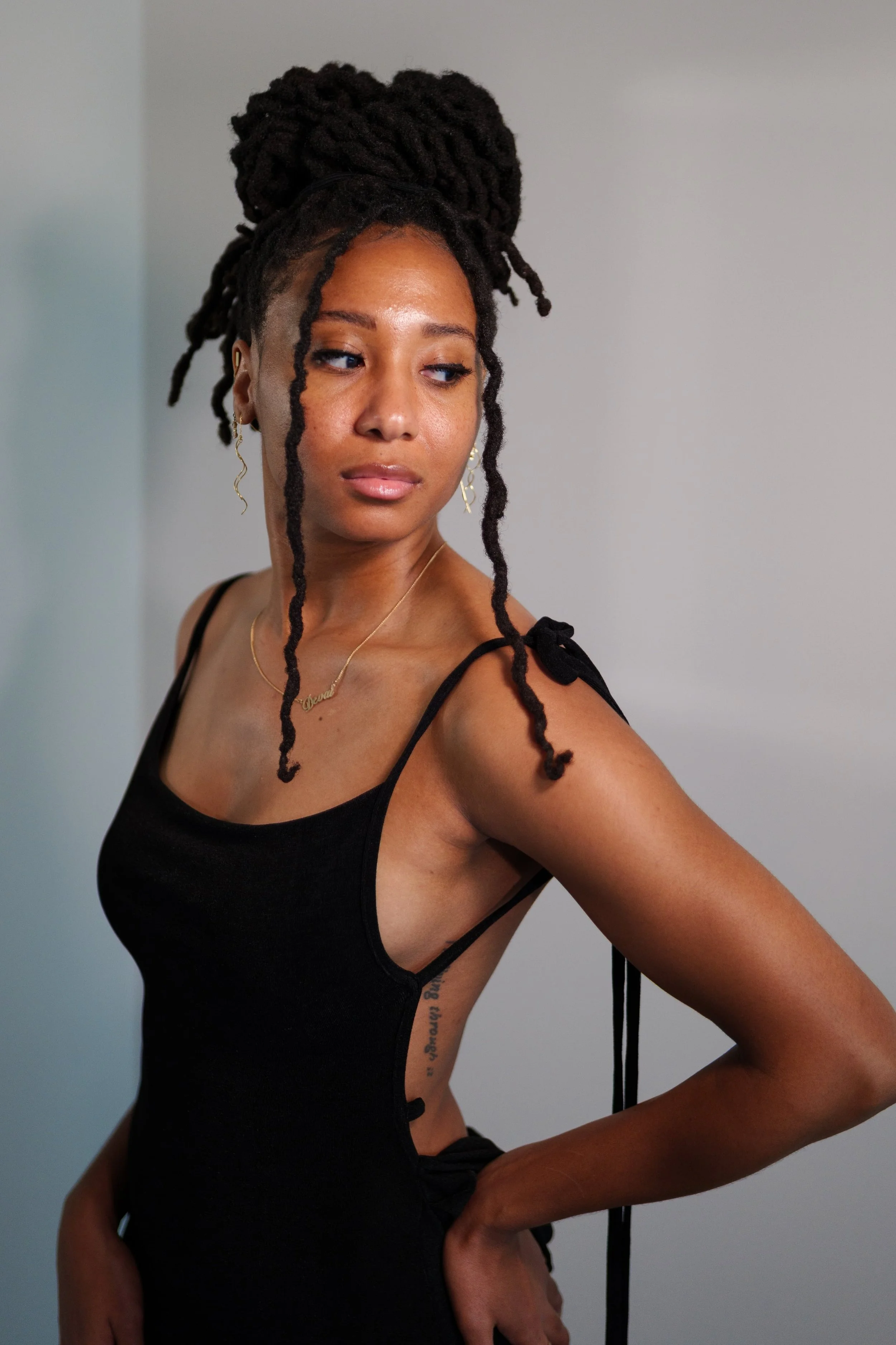 A woman with styled dreadlocks tied in a high bun, wearing a black spaghetti strap top, gold earrings, and a gold necklace, standing against a plain gray background.