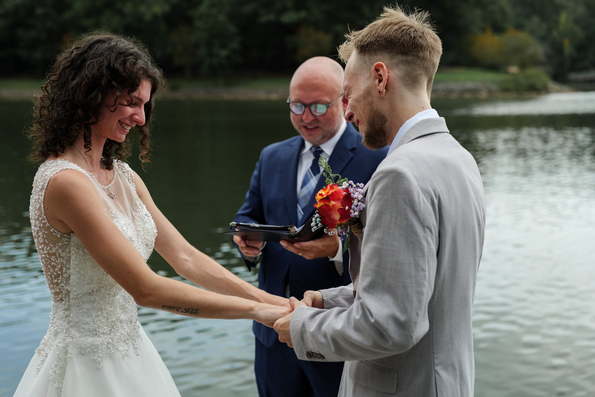 A couple getting married by a lake, holding hands and smiling at each other. An officiant stands behind them holding a book, with trees visible in the background.