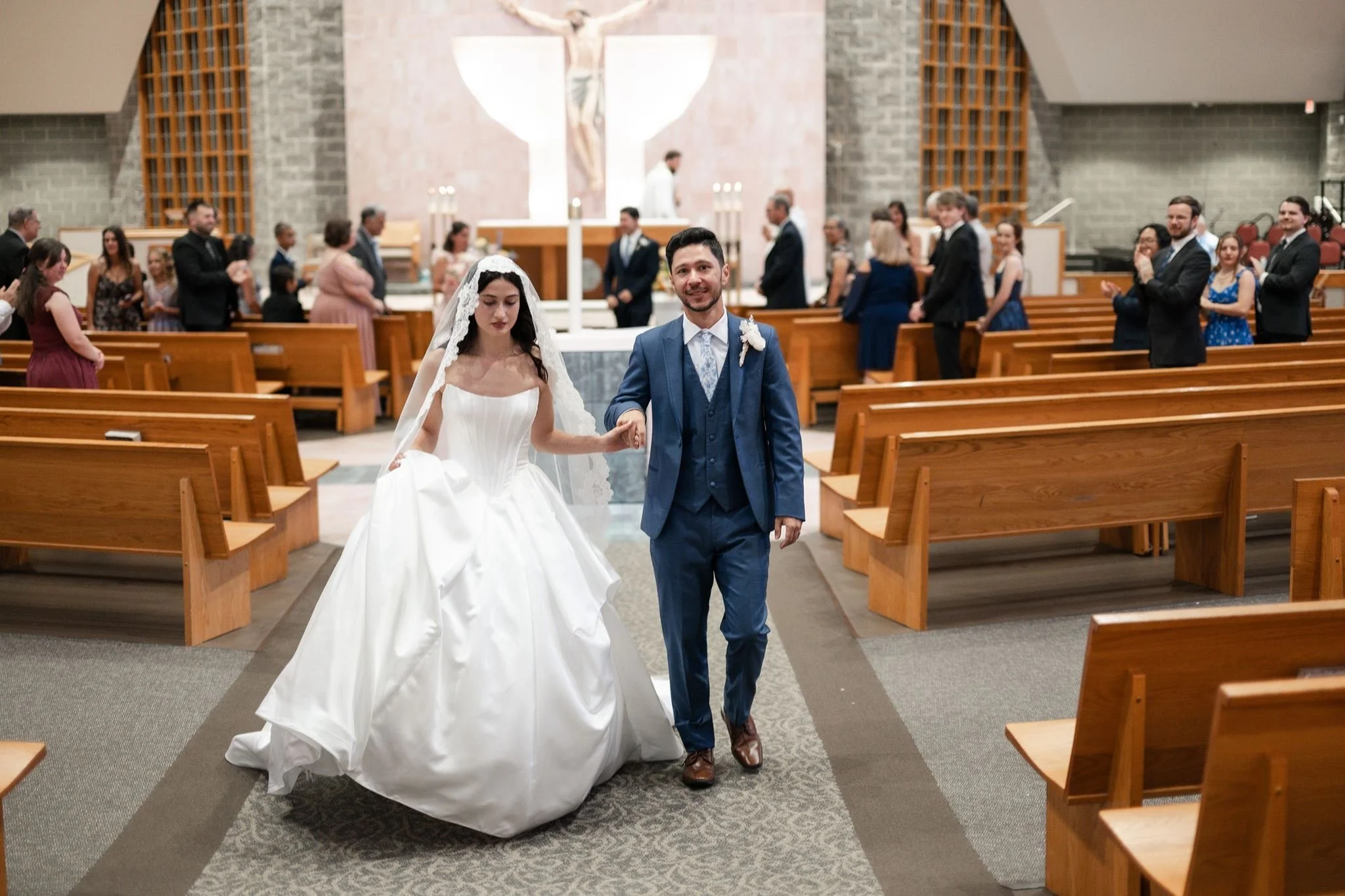 A bride and groom holding hands and walking down the aisle inside St. Mathews Catholic Church. The bride wears a white wedding gown and veil, and the groom is in a blue suit. Guests are clapping and smiling on either side of the aisle.