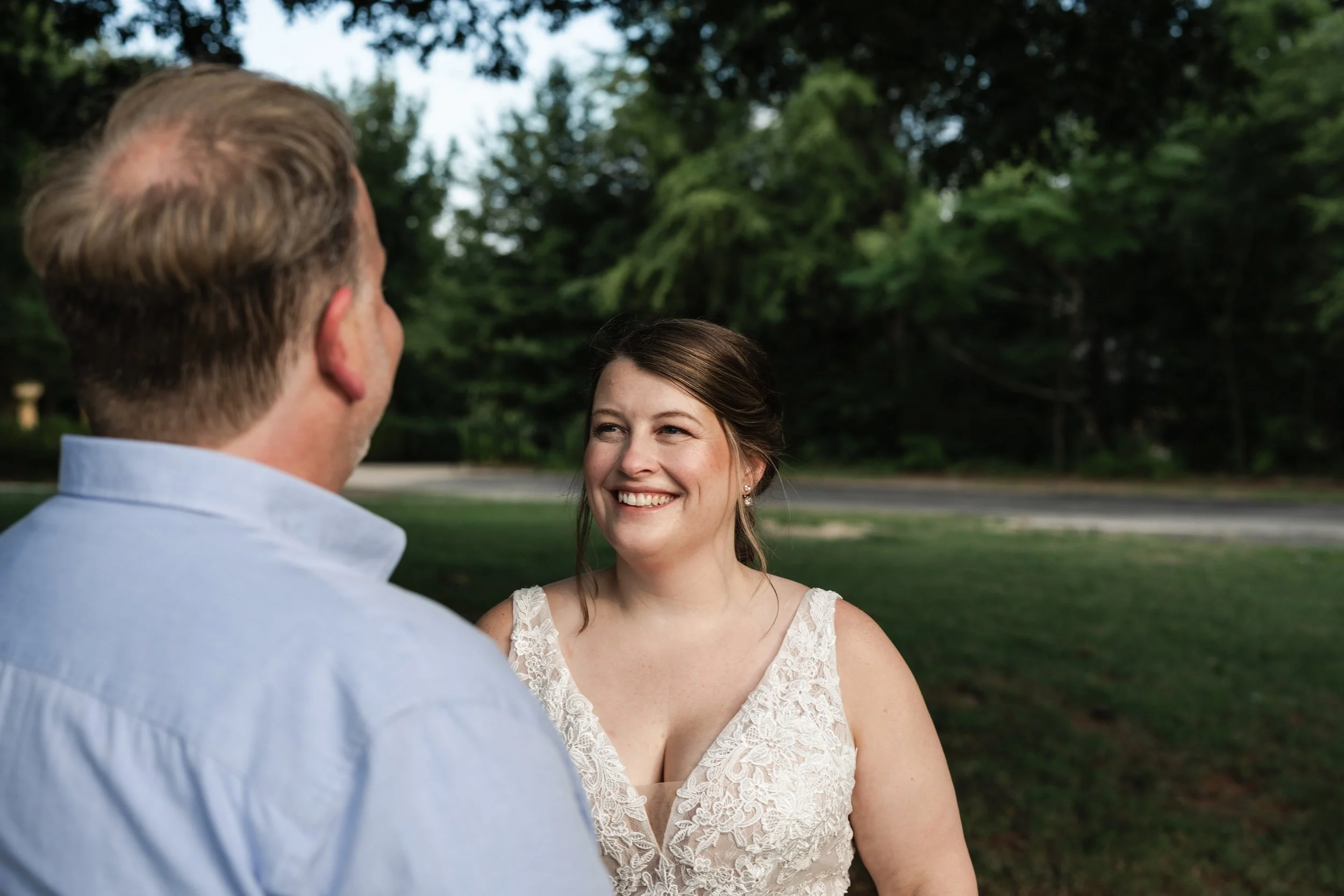 A smiling woman in a white lace dress standing outdoors, looking at a man with short, light brown hair and a light blue shirt, with a background of green trees at Daniel Stowe Botanical Gardens wedding
