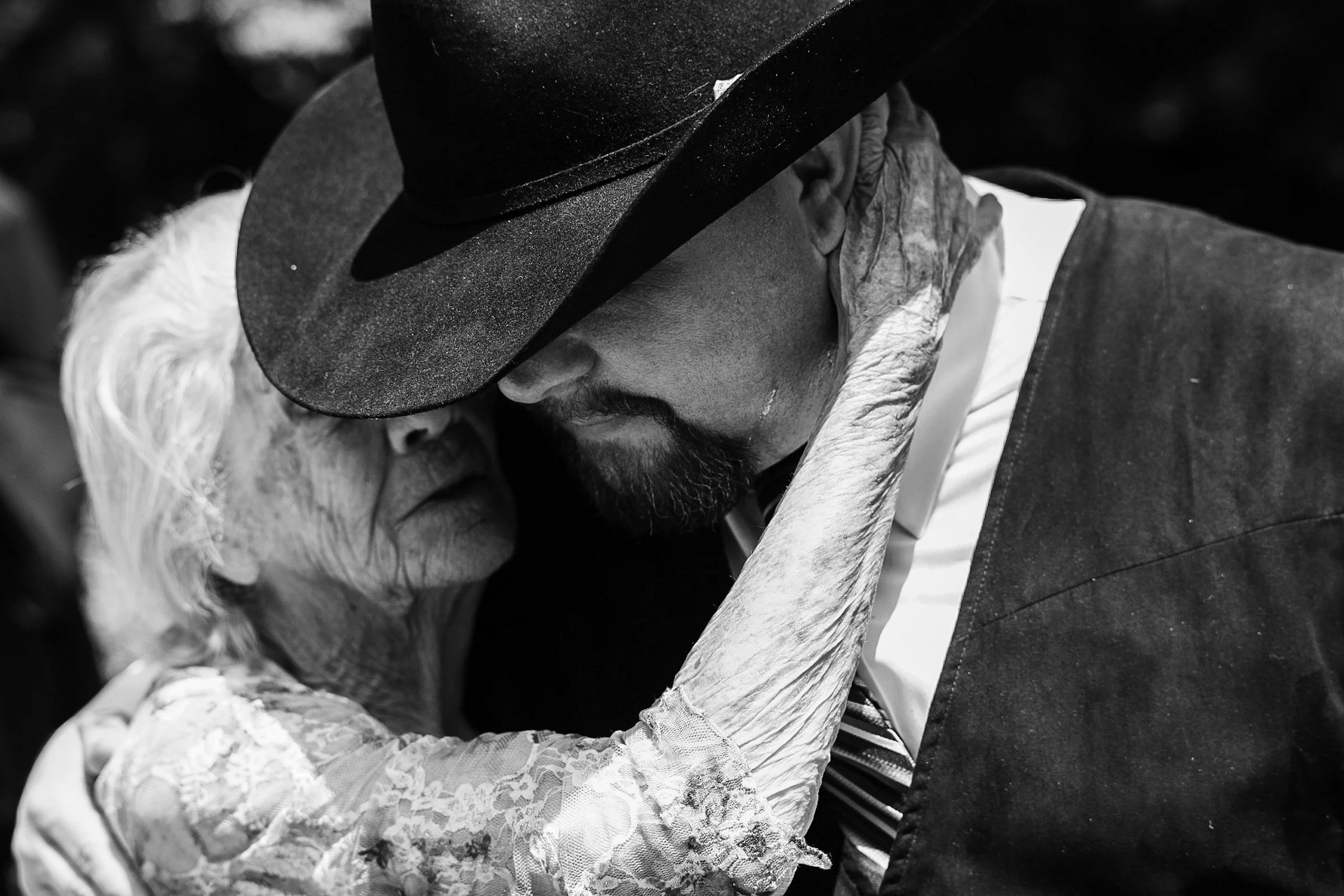 An elderly woman and a man in a cowboy hat are sharing a tender moment, touching foreheads closely.