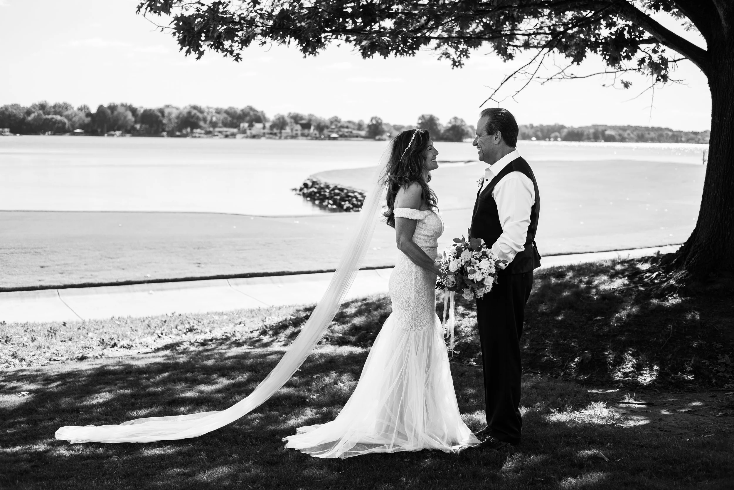 A black and white photo of a bride and groom standing under a large tree by a lake, smiling at each other, with the bride holding a bouquet of flowers.