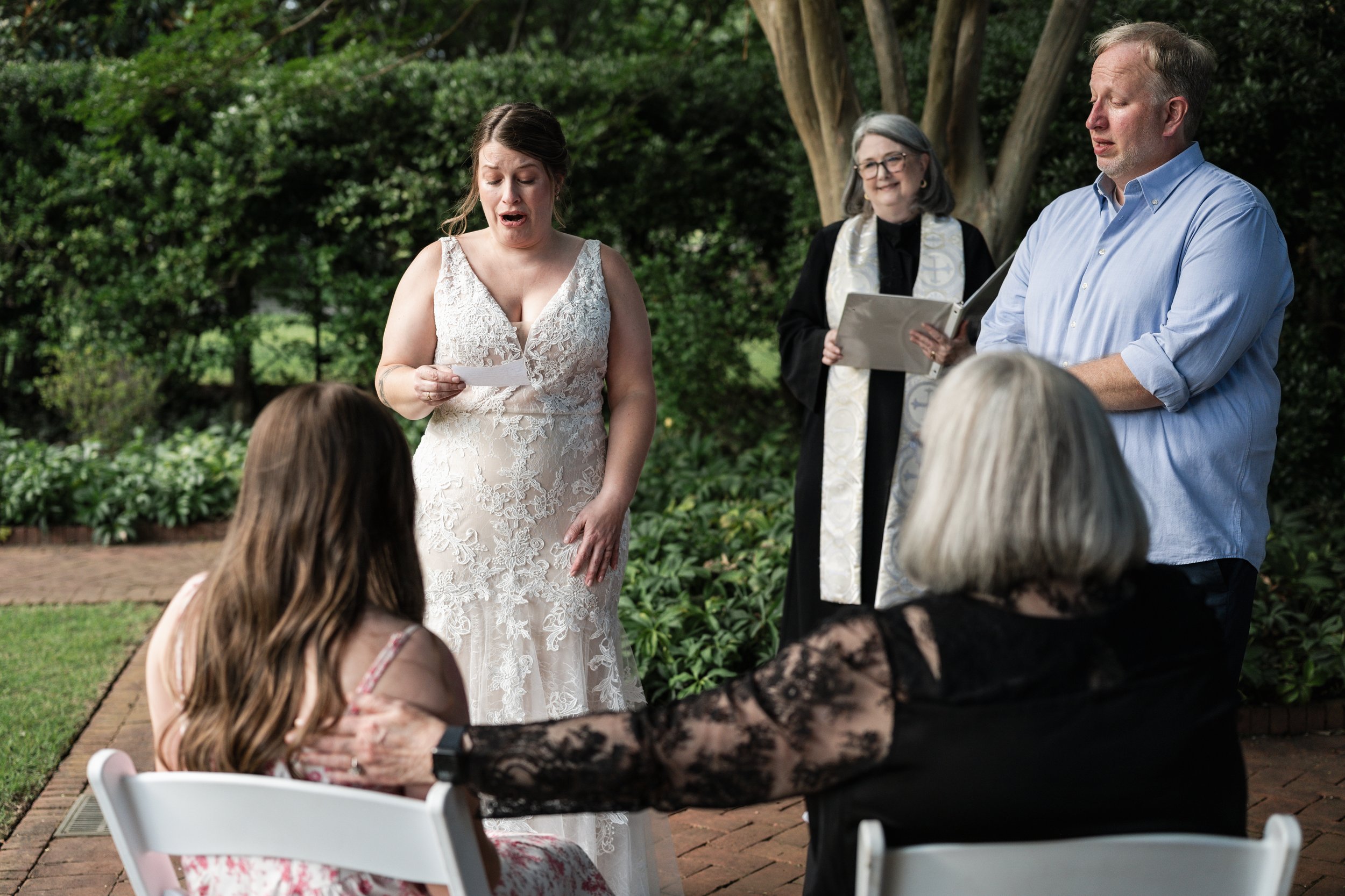 A woman in a white lace dress is reading from a piece of paper during an outdoor ceremony at Daniel Stowe Botanical Gardens, with a man and a woman officiating and guests seated nearby.
