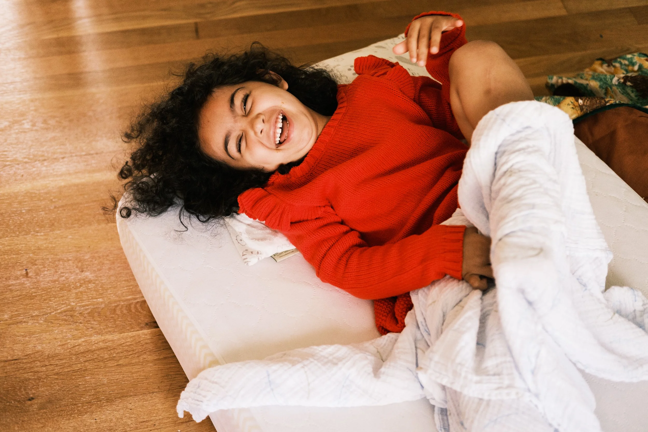 A young woman with curly black hair and light brown skin, lying on a bed, smiling and laughing, wearing a red sweater, with a white blanket partially covering her legs.