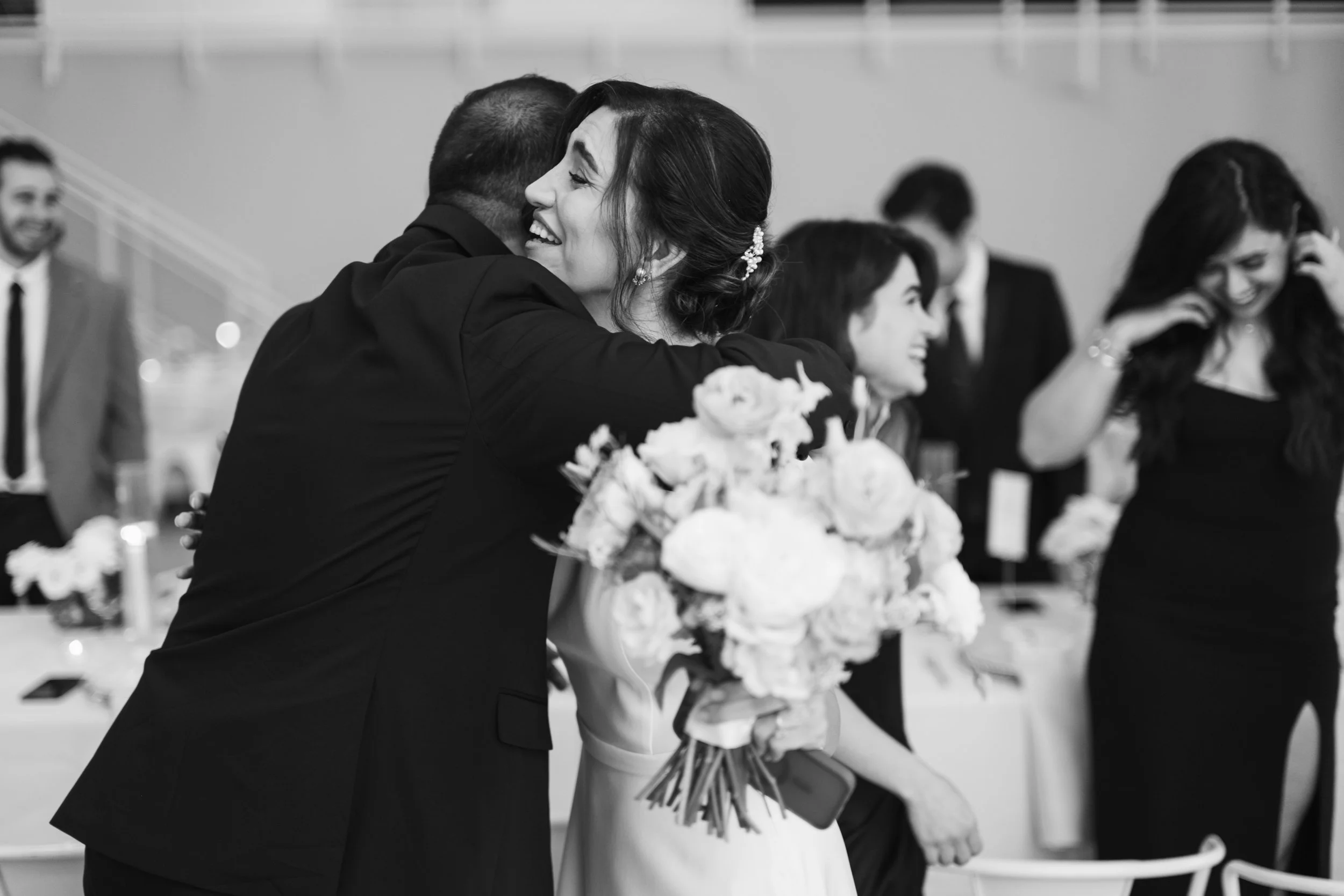 Black and white photo of a bride in a white dress holding a bouquet smiling while hugging a family member during her wedding in NoDa, Charlotte.
