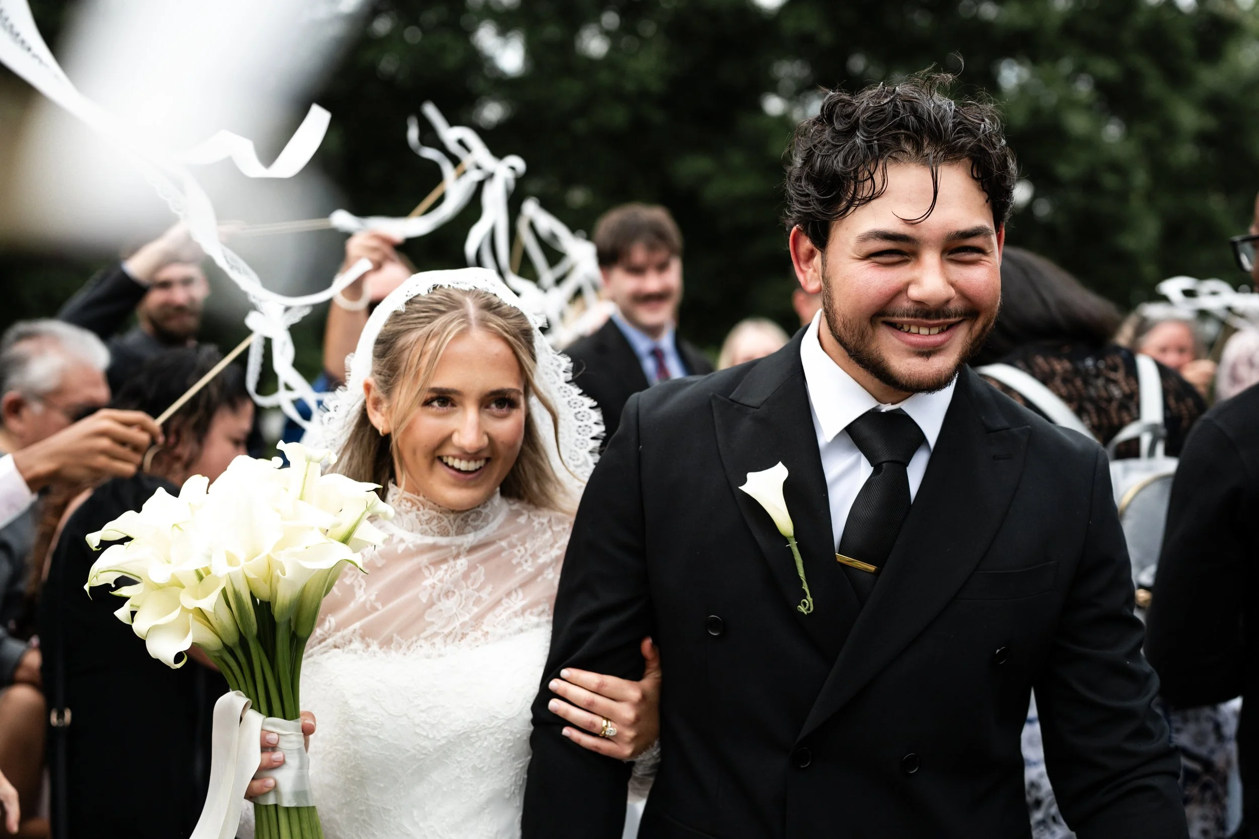 A smiling bride and groom walking outside St. Mary's Chapel on their wedding day, surrounded by guests. The bride is holding a bouquet of white calla lilies, and the groom is wearing a black suit with a white shirt and black tie.