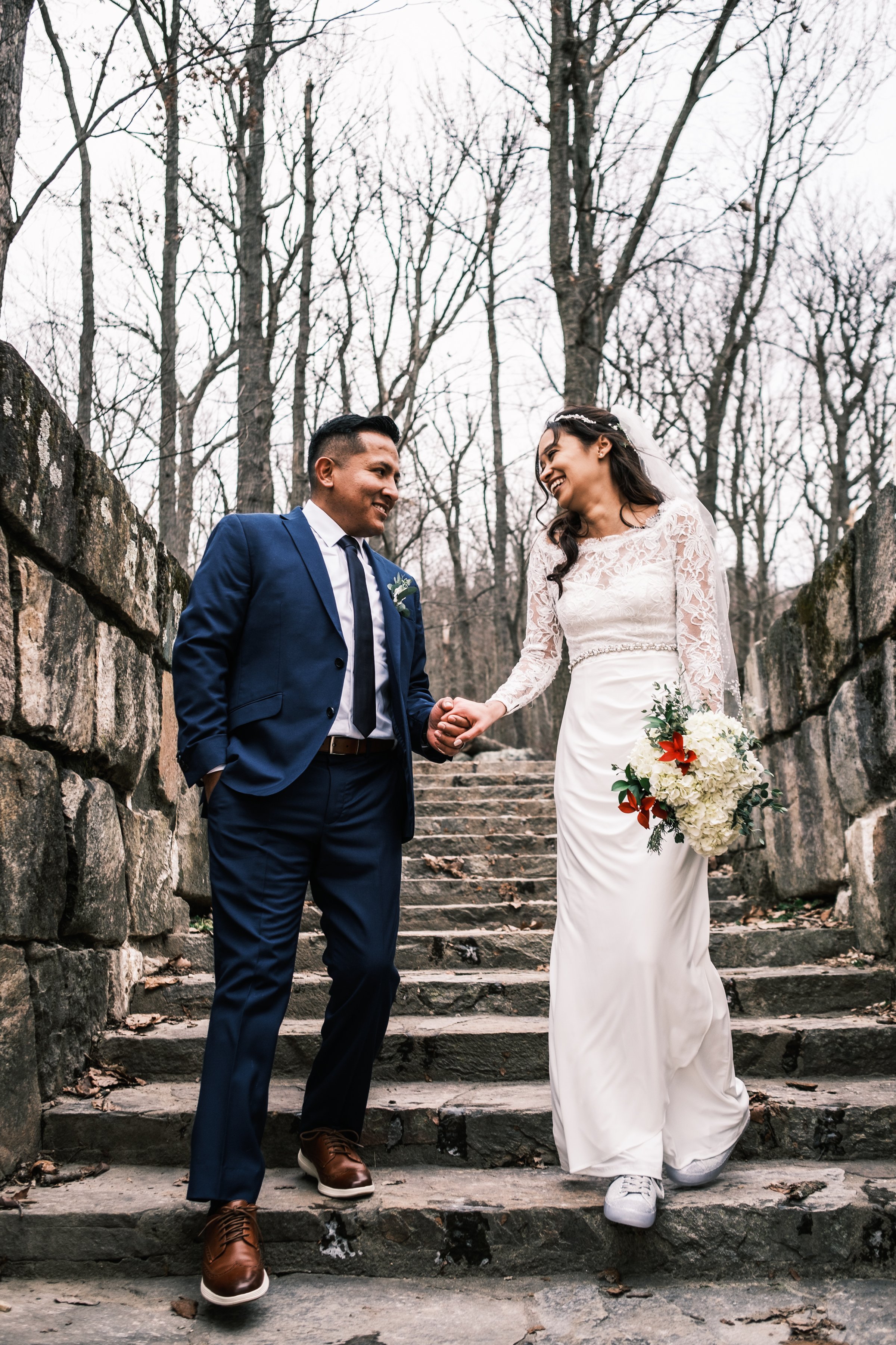 A bride in a white lace wedding dress and a groom in a blue suit holding hands and smiling on stone steps outdoors with leafless trees in the background.