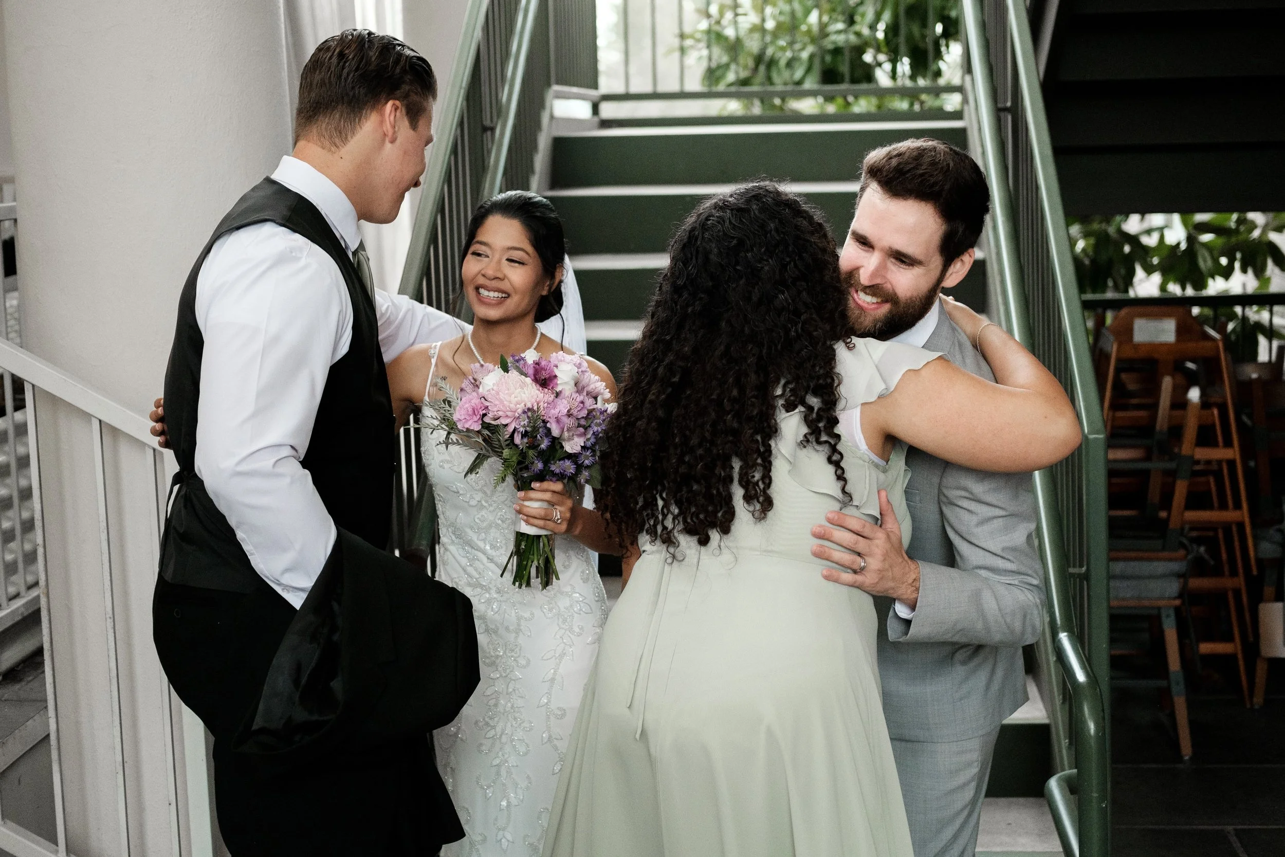 People hugging and smiling in a wedding celebration indoors, with a bride holding a bouquet of pink and purple flowers in the background.