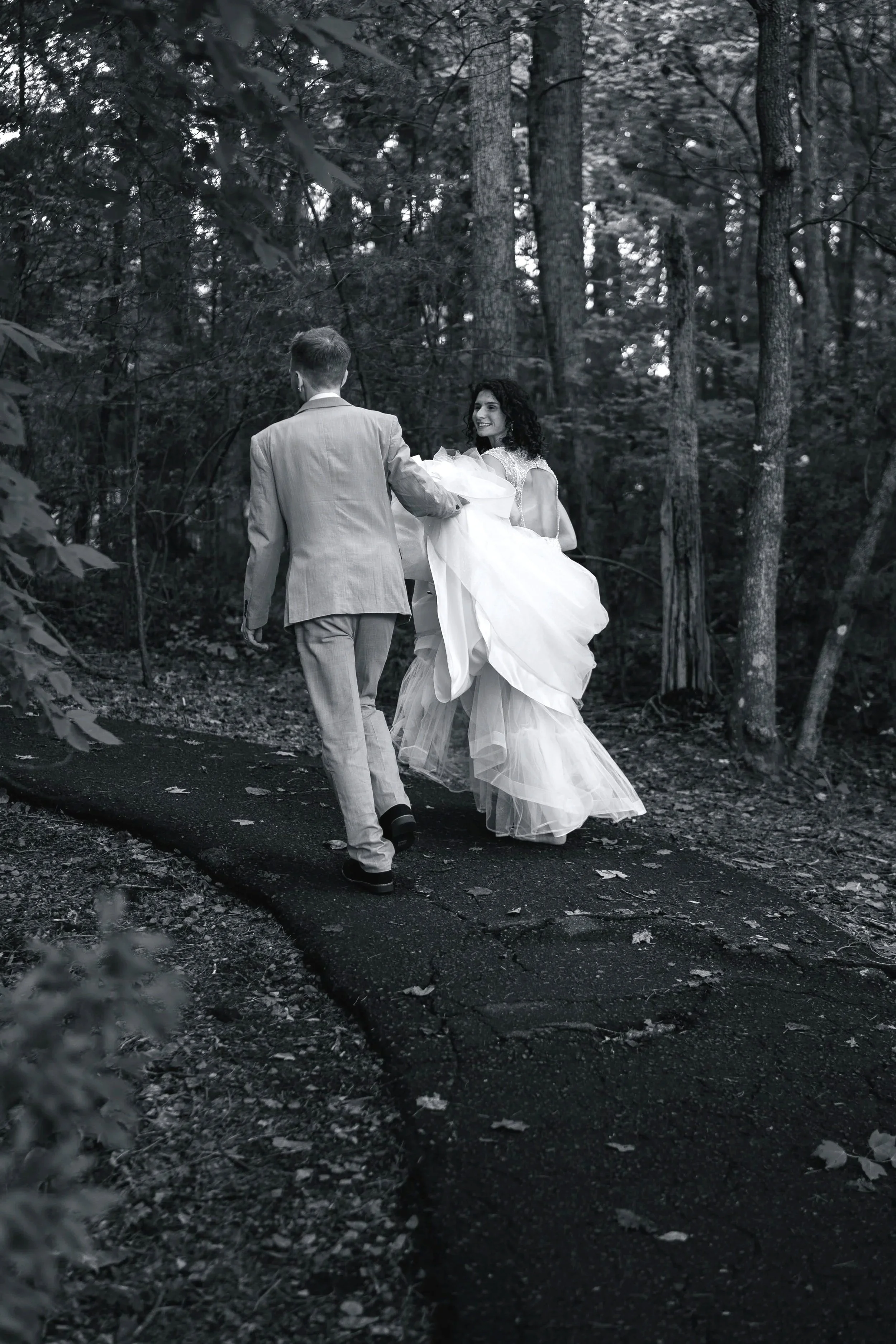 A black and white photo of a bride and groom walking through a forest. The bride is wearing a wedding gown and holding the groom's hand, smiling at him. The groom is wearing a light-colored suit, with his back to the camera. The scene appears romantic and intimate.