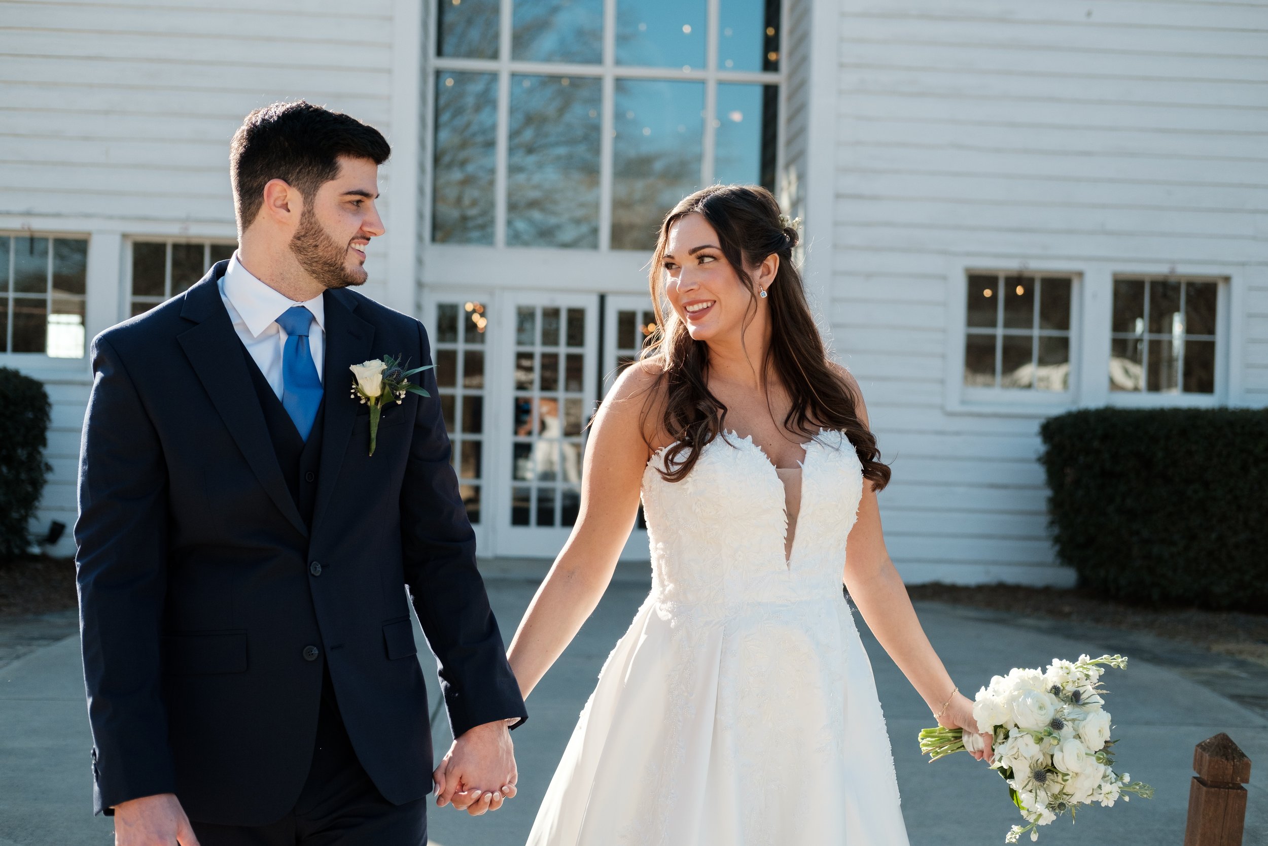 A bride and groom holding hands outside a white building with large windows, smiling at each other, during their wedding at Founders Dairy Barn in Fort Mill, SC.