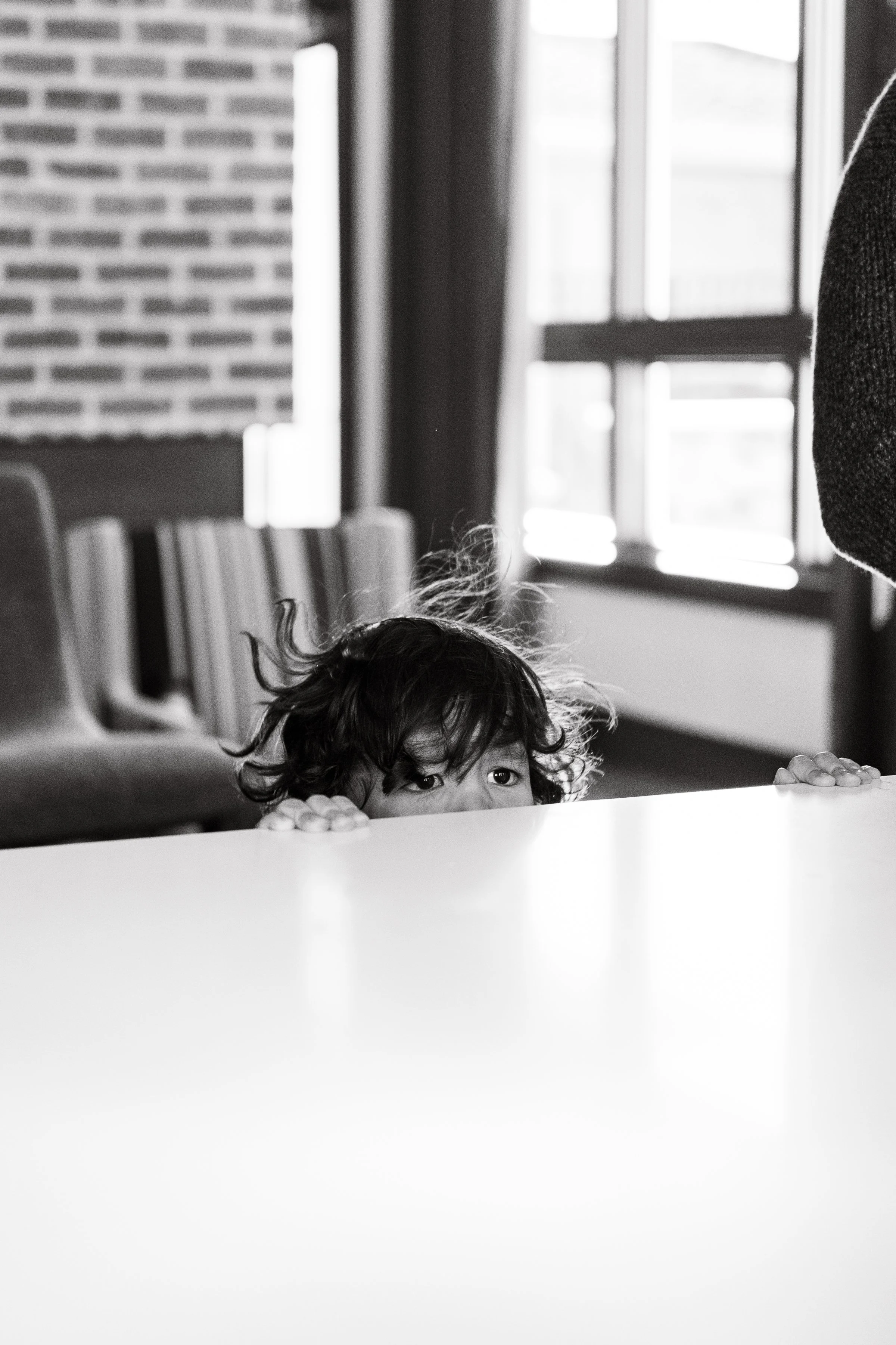 A young child with curly hair looking over the edge of a surface, with a background featuring a brick wall and a window.