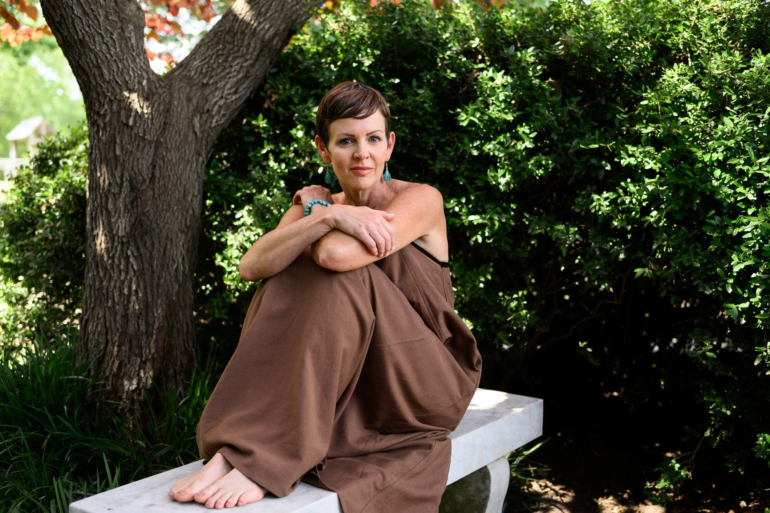 A woman with short brown hair sitting outdoors on a stone bench, surrounded by green bushes and trees, wearing a brown dress and turquoise jewelry.