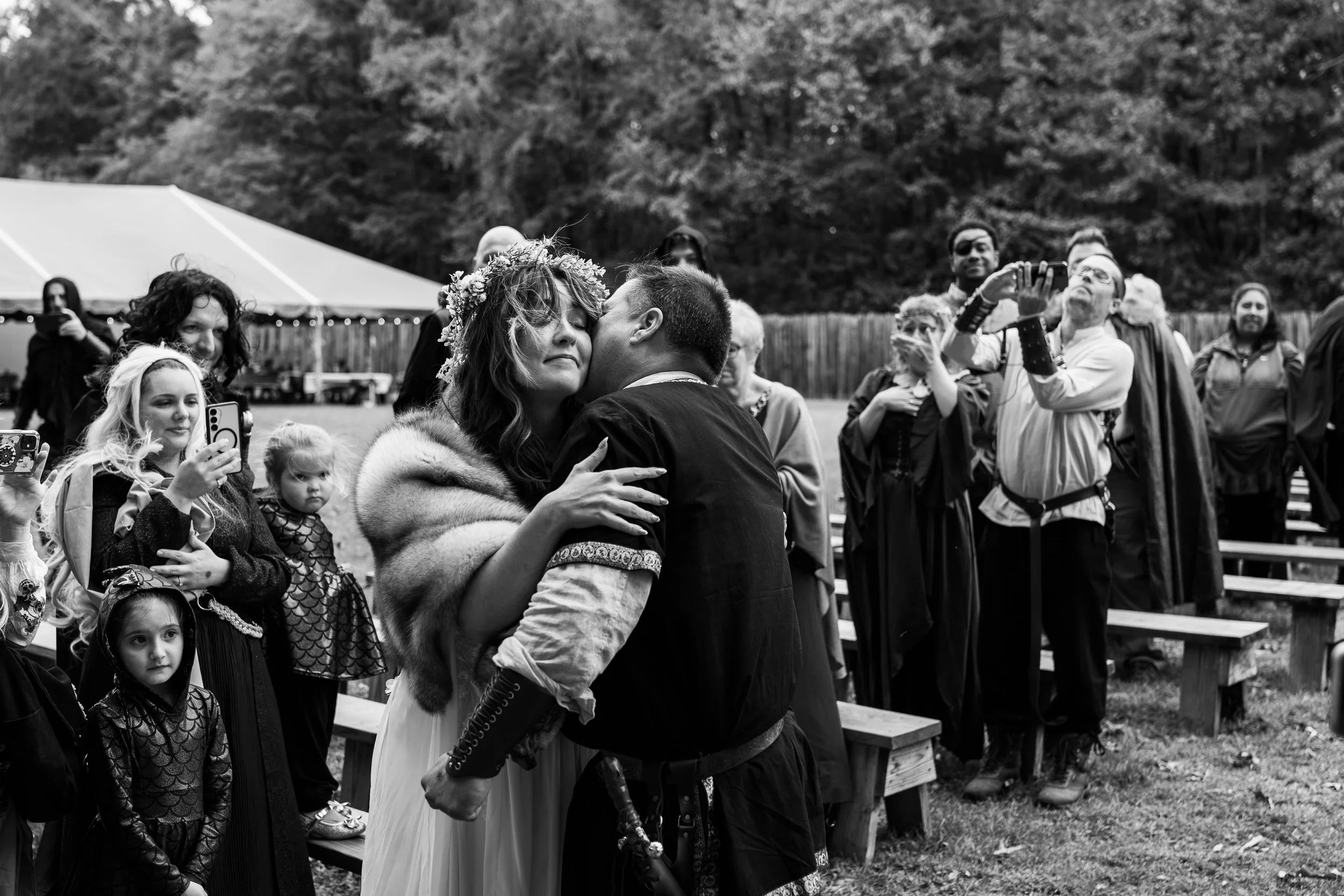 A group of people gathered outdoors at a renaissance wedding in Charlotte , with a couple embracing in the center as others watch and take photos. Many are dressed in fantasy or medieval costumes.