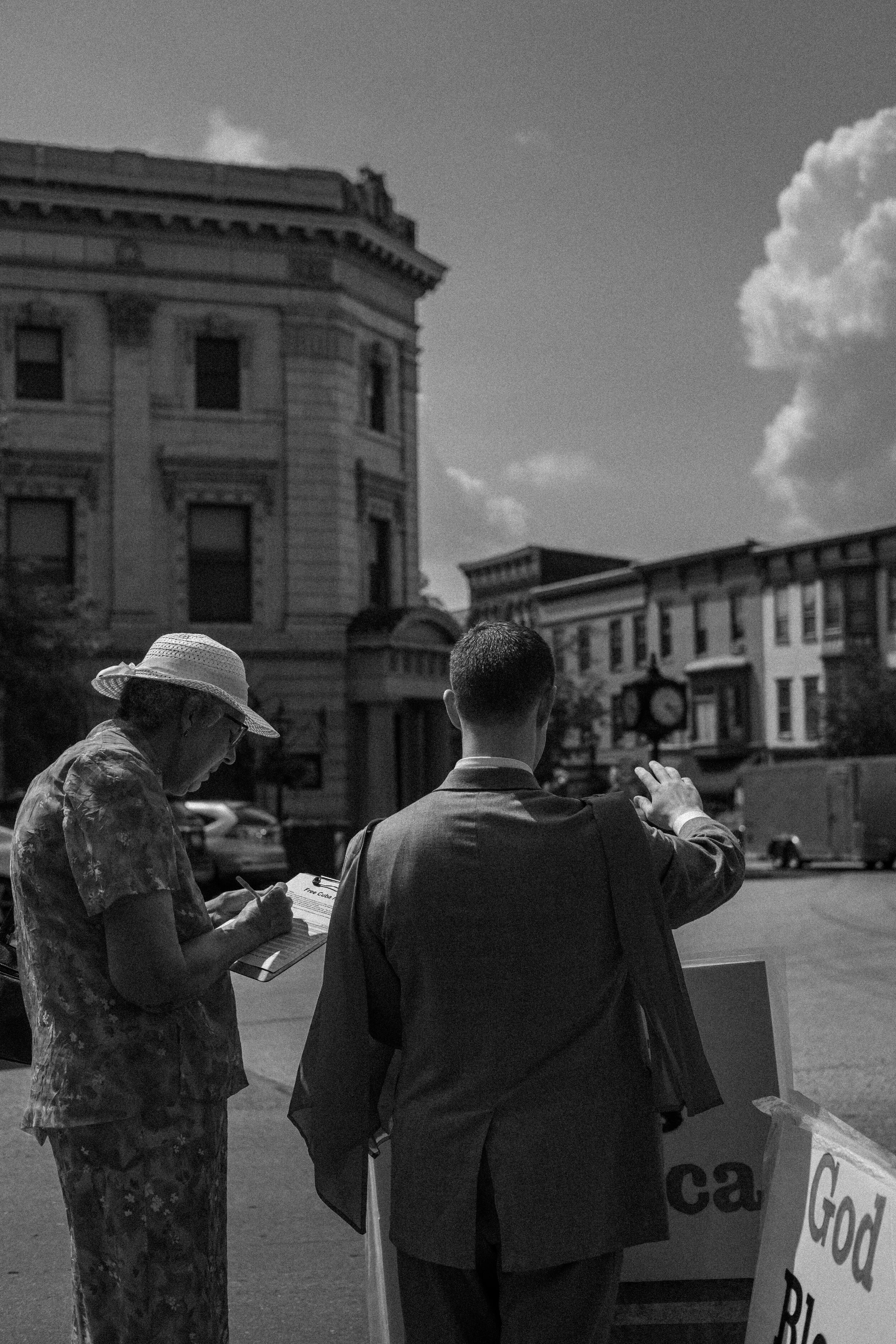 Black and white photo of two people engaged in a discussion on a city street. One person, wearing a hat, is writing on a clipboard. The other person, in a suit, gestures towards the street. An old building and a clock are in the background, with a pa