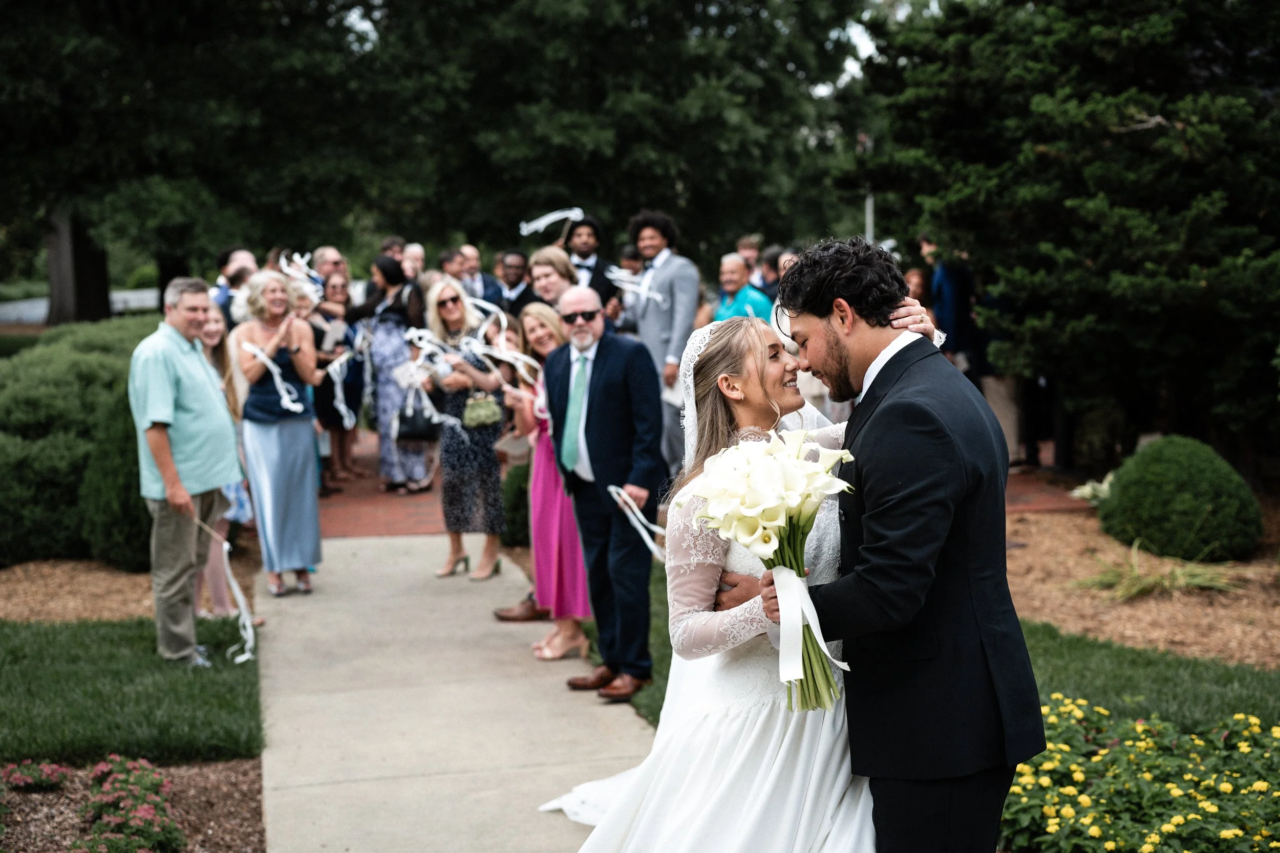 A bride and groom are about to kiss at their outdoor wedding ceremony at St. Mary's Chapel in Charlotte, with guests in the background watching and smiling.