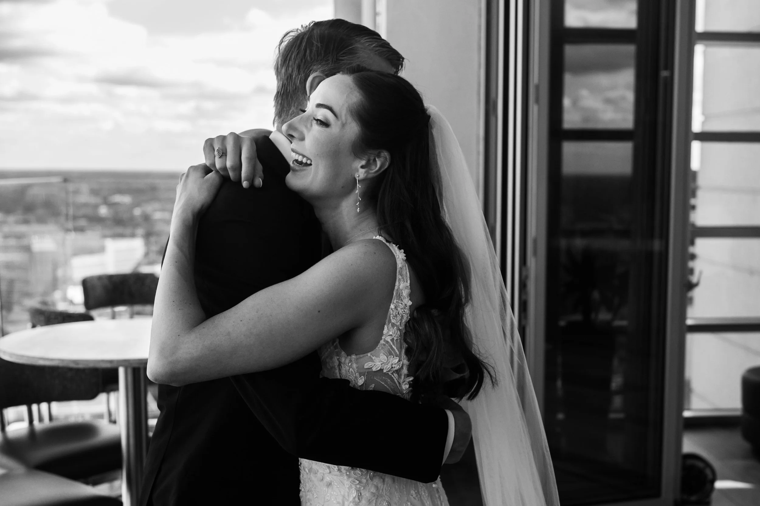 A bride and her father embrace during their wedding photography at Nuvole Rooftop, smiling and holding each other closely indoors with large windows in the background.