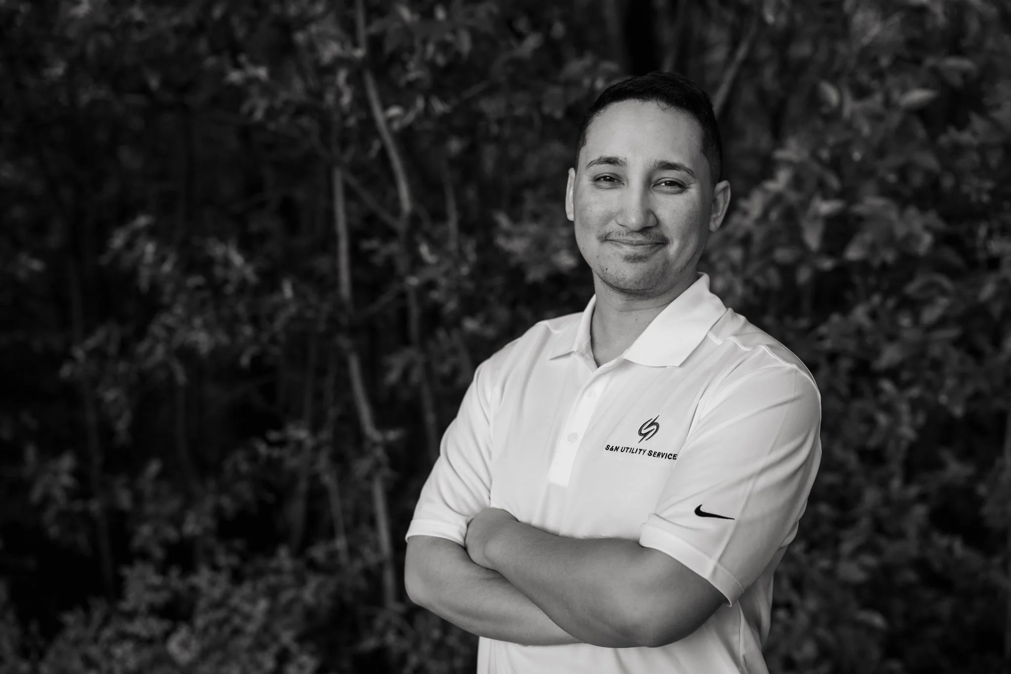 A man in a white collared shirt with a logo and the text 'S&M Utility Service' is standing outdoors in front of a background of leaves. He has short dark hair, a slight smile, and his arms crossed.