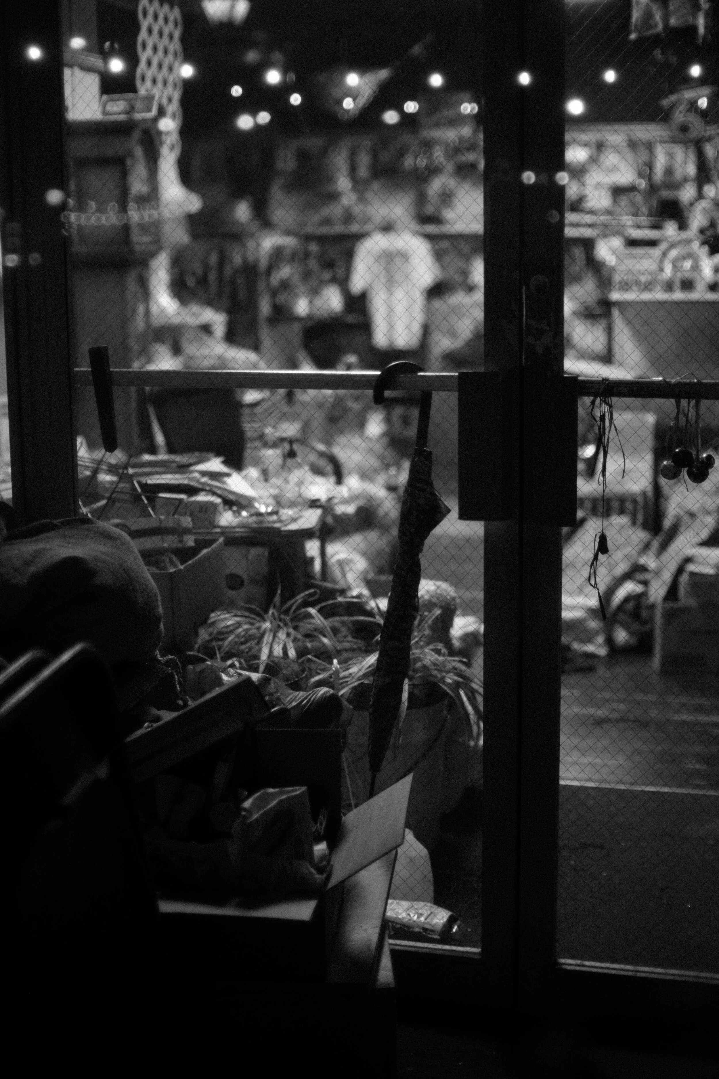 Black and white interior of a cluttered shop seen through a glass door with a netting design. Items include boxes, an umbrella, and hanging decorations. Blurred lights and clothing hang in the background.