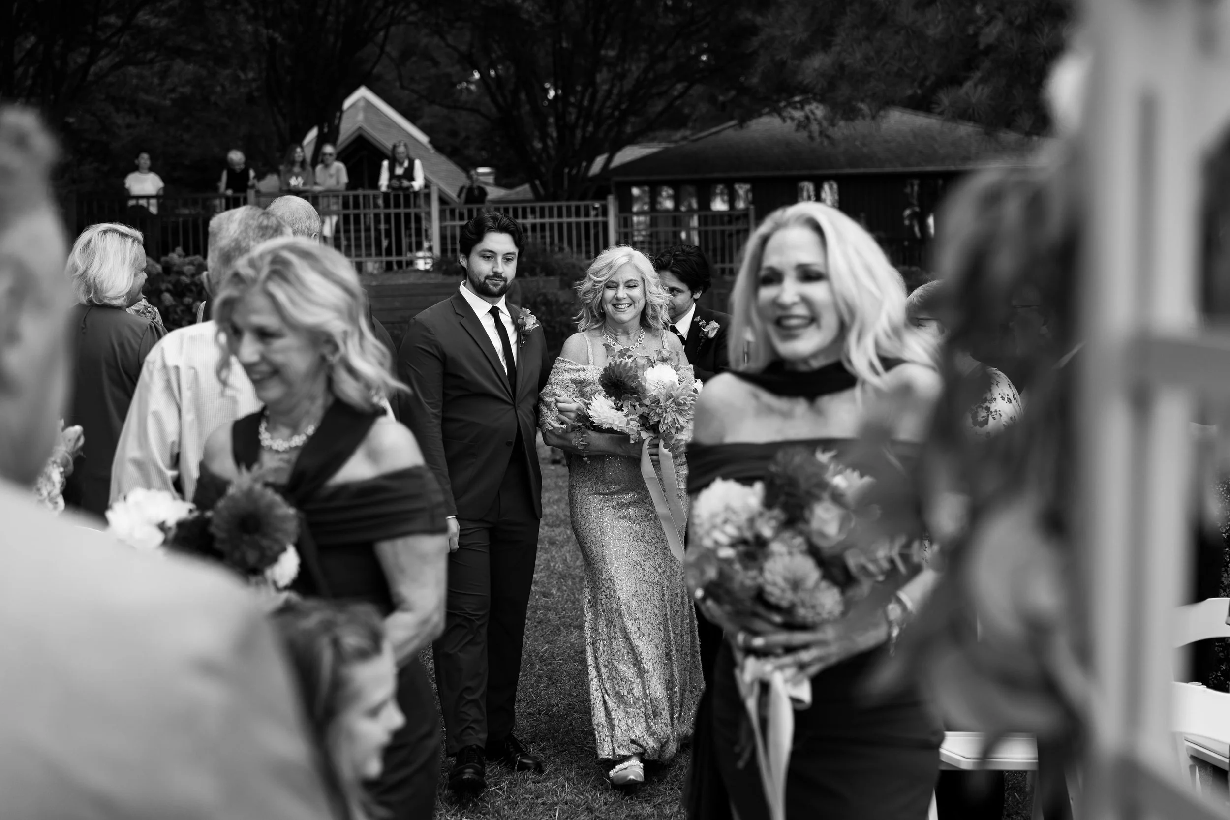 People attending a wedding ceremony outdoors, with a woman in a glittery dress and a man in a suit as central figures, surrounded by guests holding flowers, and trees and a pavilion in the background at Jetty Park, Lake Norman