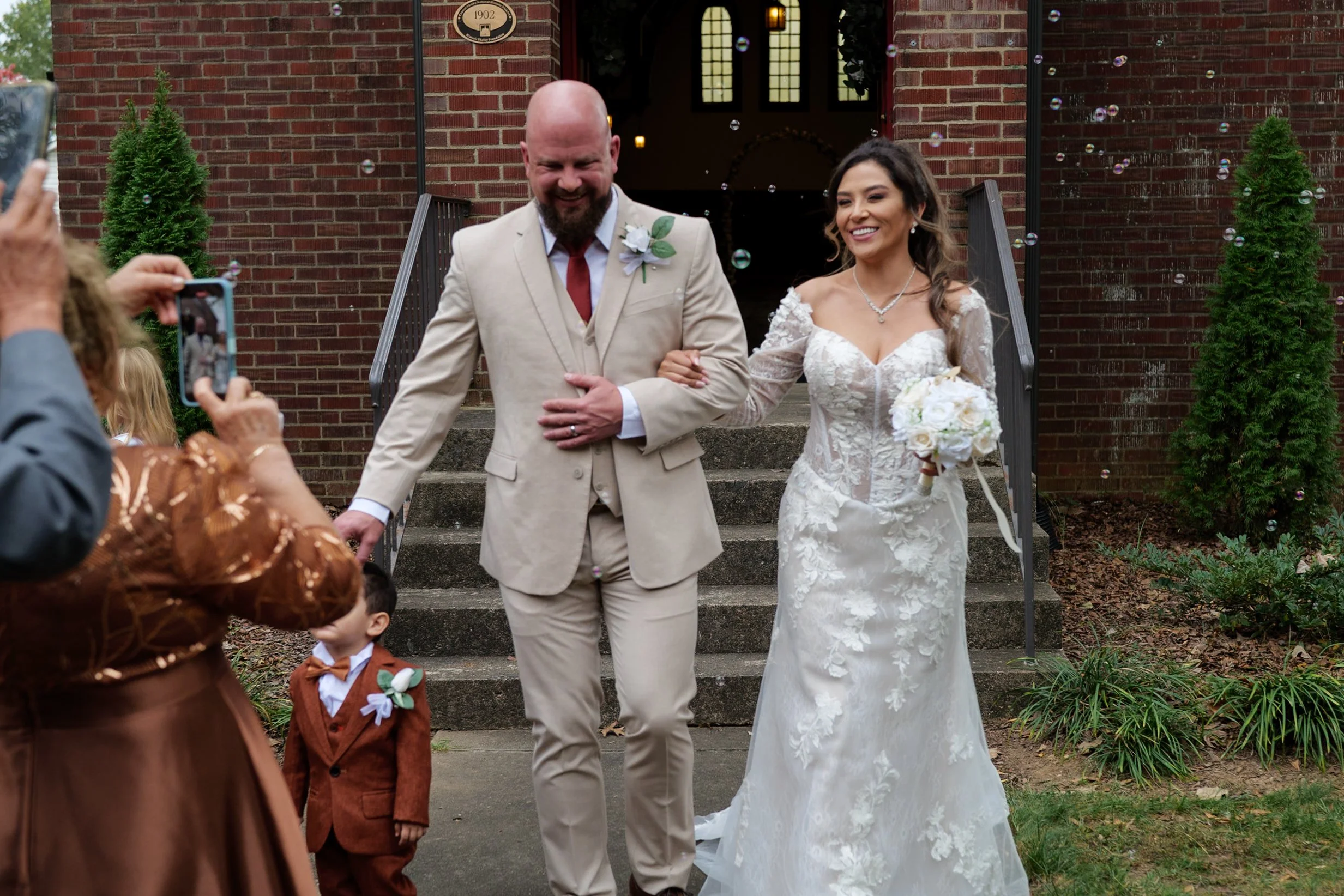 Happy bride and groom descending steps outside a brick church, holding hands, surrounded by family and friends. The bride wears a white lace wedding gown and holds a bouquet, and the groom wears a light-colored suit with a boutonniere. Guests are tak