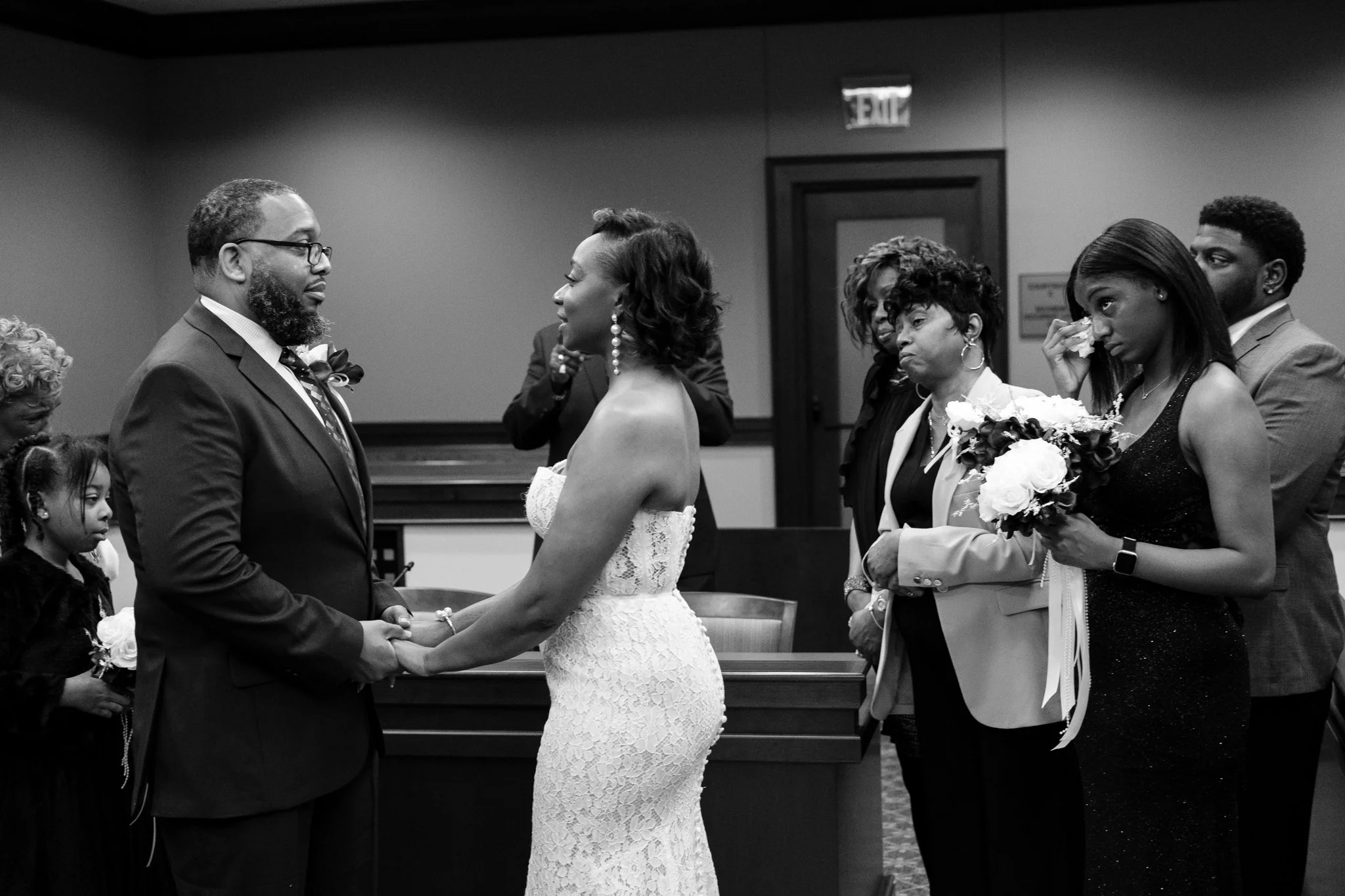 A black and white photo of a wedding ceremony with a couple holding hands and facing each other, surrounded by guests in formal attire. The bride is in a strapless lace wedding dress, and the groom is in a suit with glasses, both looking at each othe