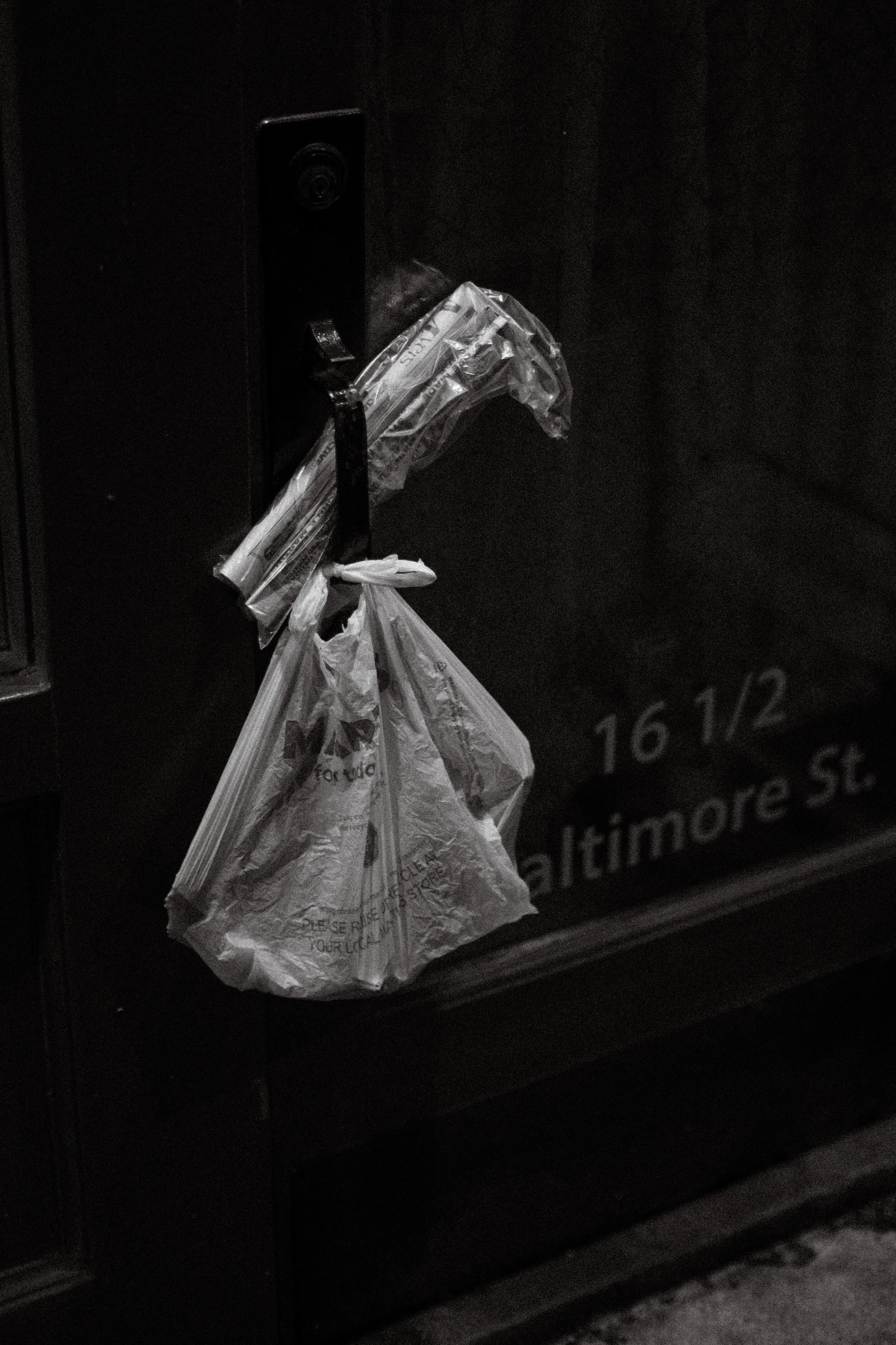 Black and white image of a plastic bag and newspapers hung on a door handle with visible text reading "16 1/2 Baltimore St." on the door.