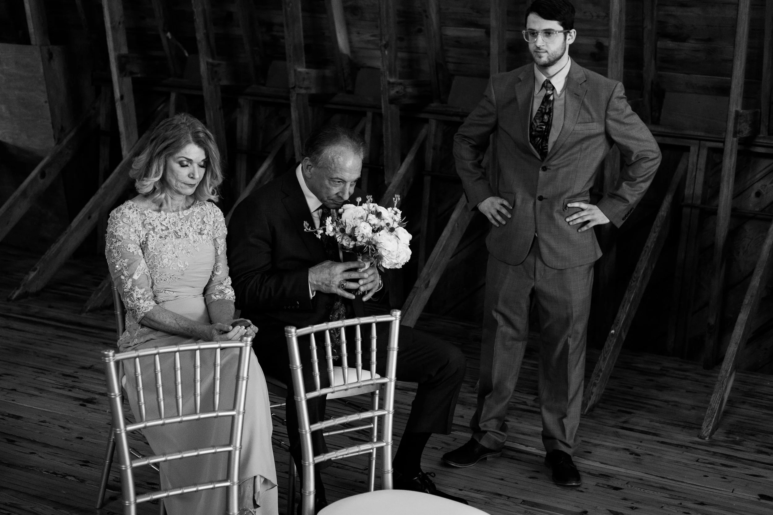 A black and white photo of three people in a solemn moment; a woman, an older man holding a bouquet of flowers, and a younger man standing with hands on hips, inside a wooden room, possibly during a ceremony.