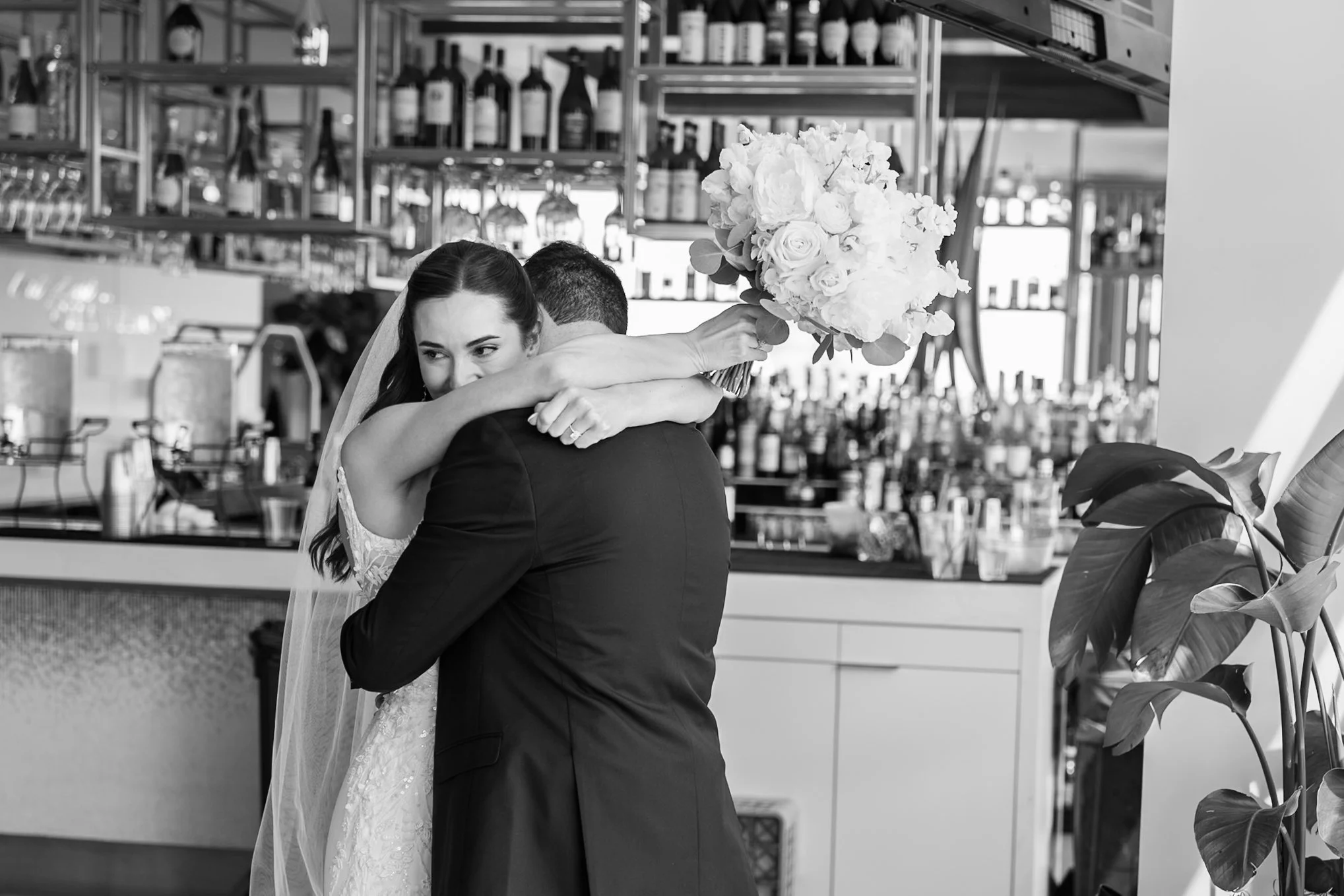 A bride and groom embrace in a restaurant or bar with shelves of bottles and glasses behind them, and a large bouquet of flowers visible.