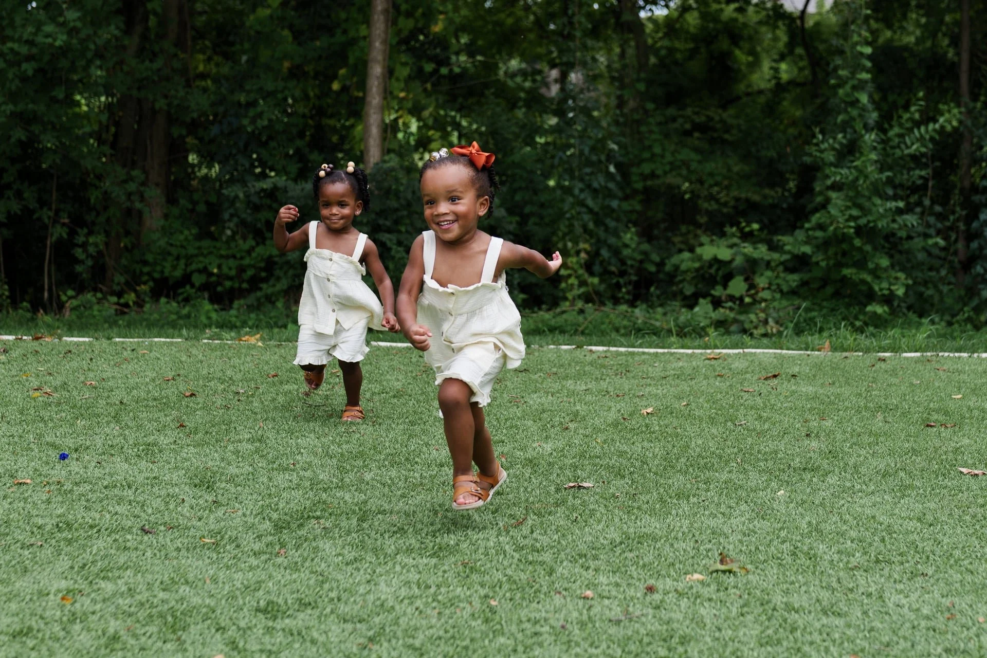 Two young twin girls running happily on a grassy lawn with a wooded background during a family photography session in Pineville, NC.