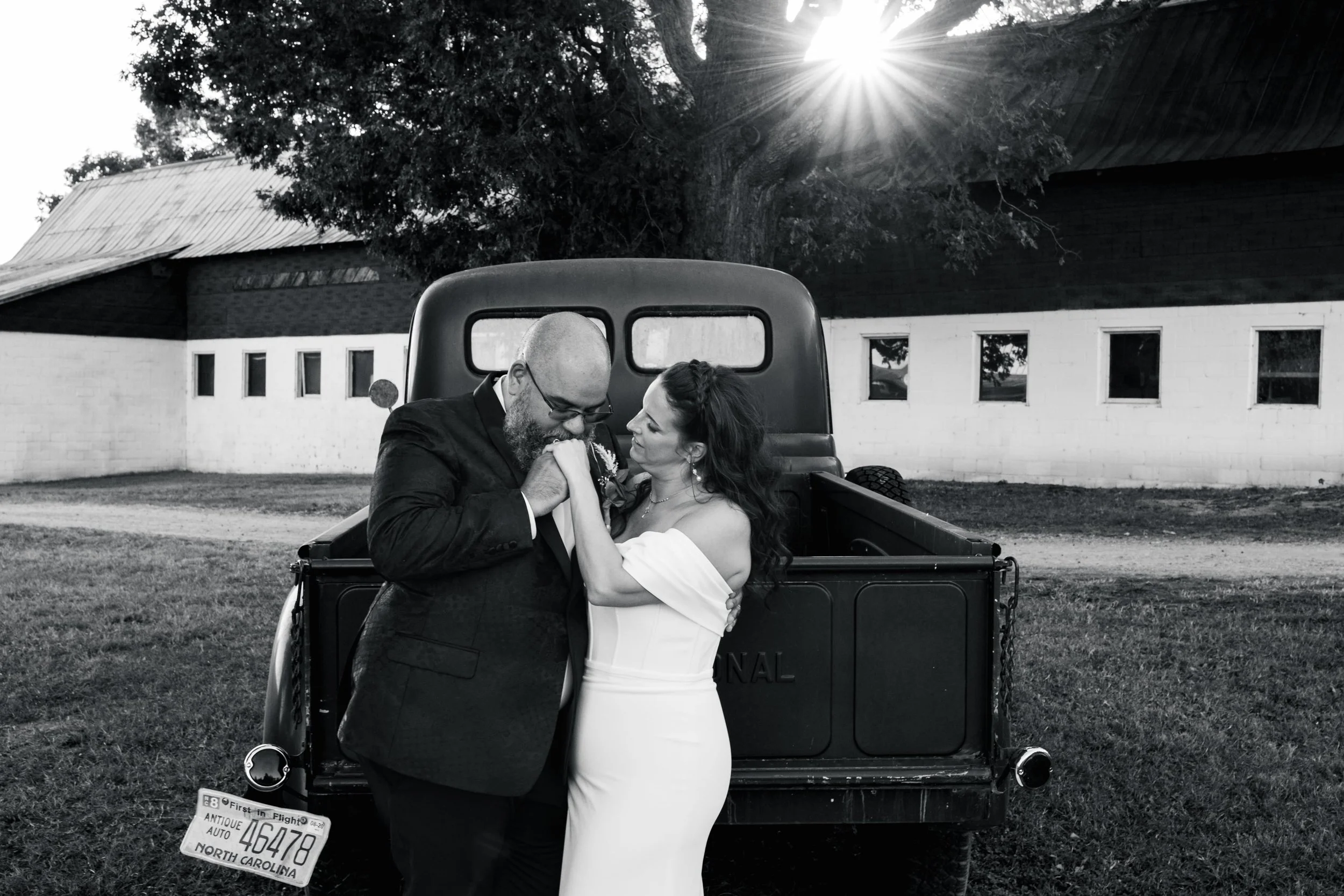 Black and white photo of a couple in wedding attire embracing in front of a vintage truck with North Carolina license plate, outside barn and trees, sun shining through branches.