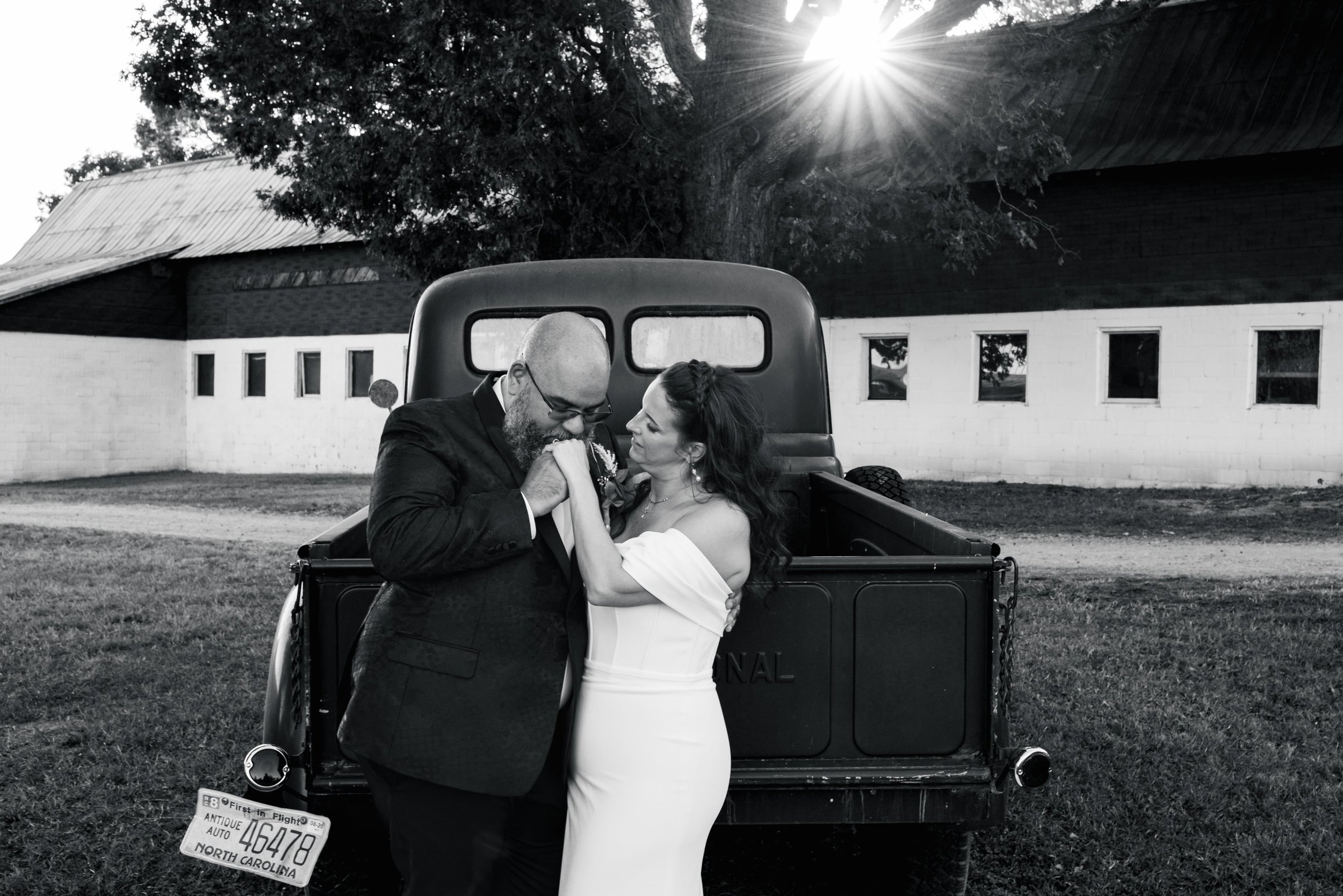 Black and white photo of a couple in wedding attire embracing in front of a vintage truck with North Carolina license plate, outside barn and trees, sun shining through branches.
