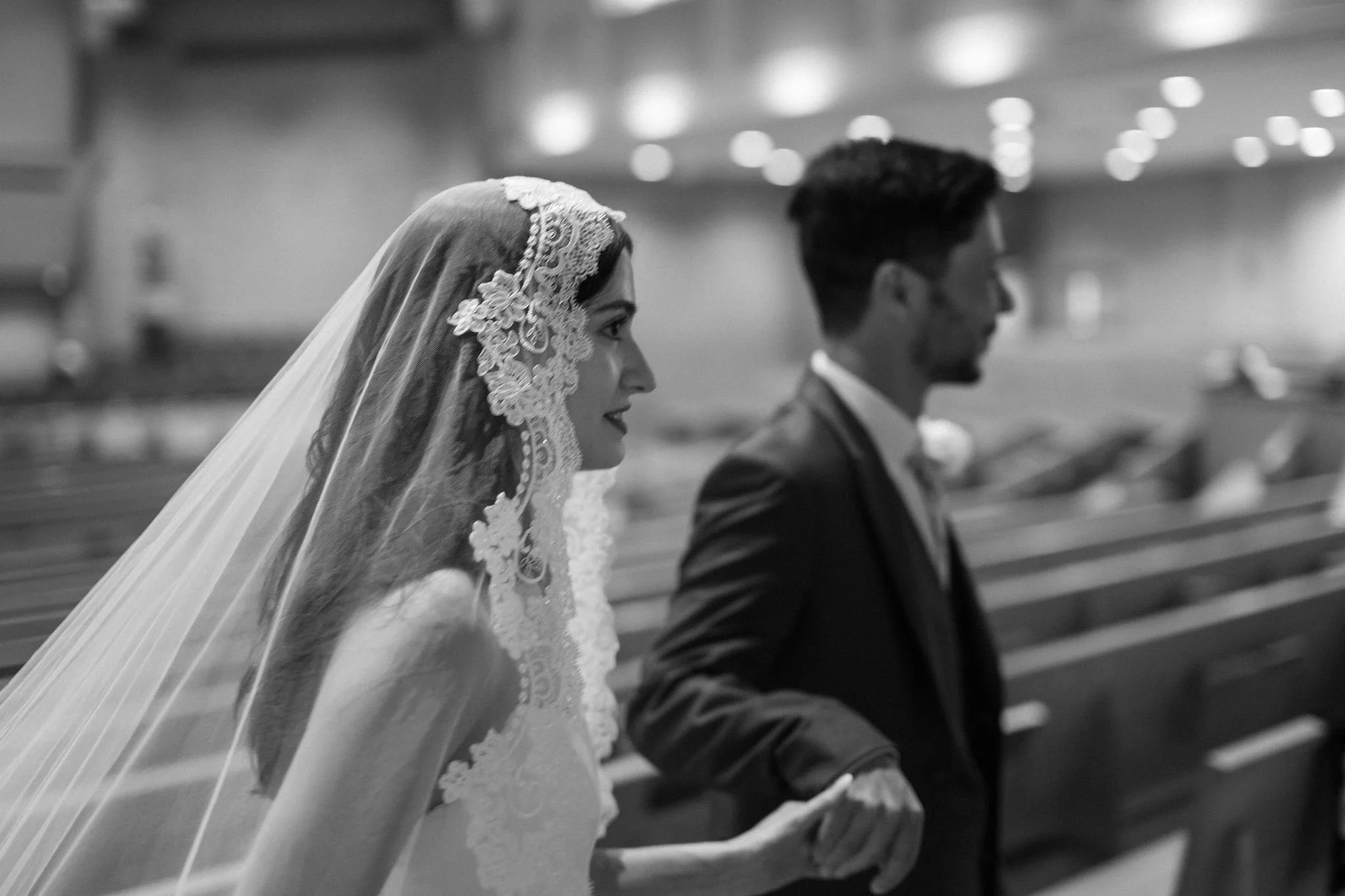 A bride and groom holding hands during their wedding ceremony inside St. Mathew Catholic church, with a blurred background of church lights.