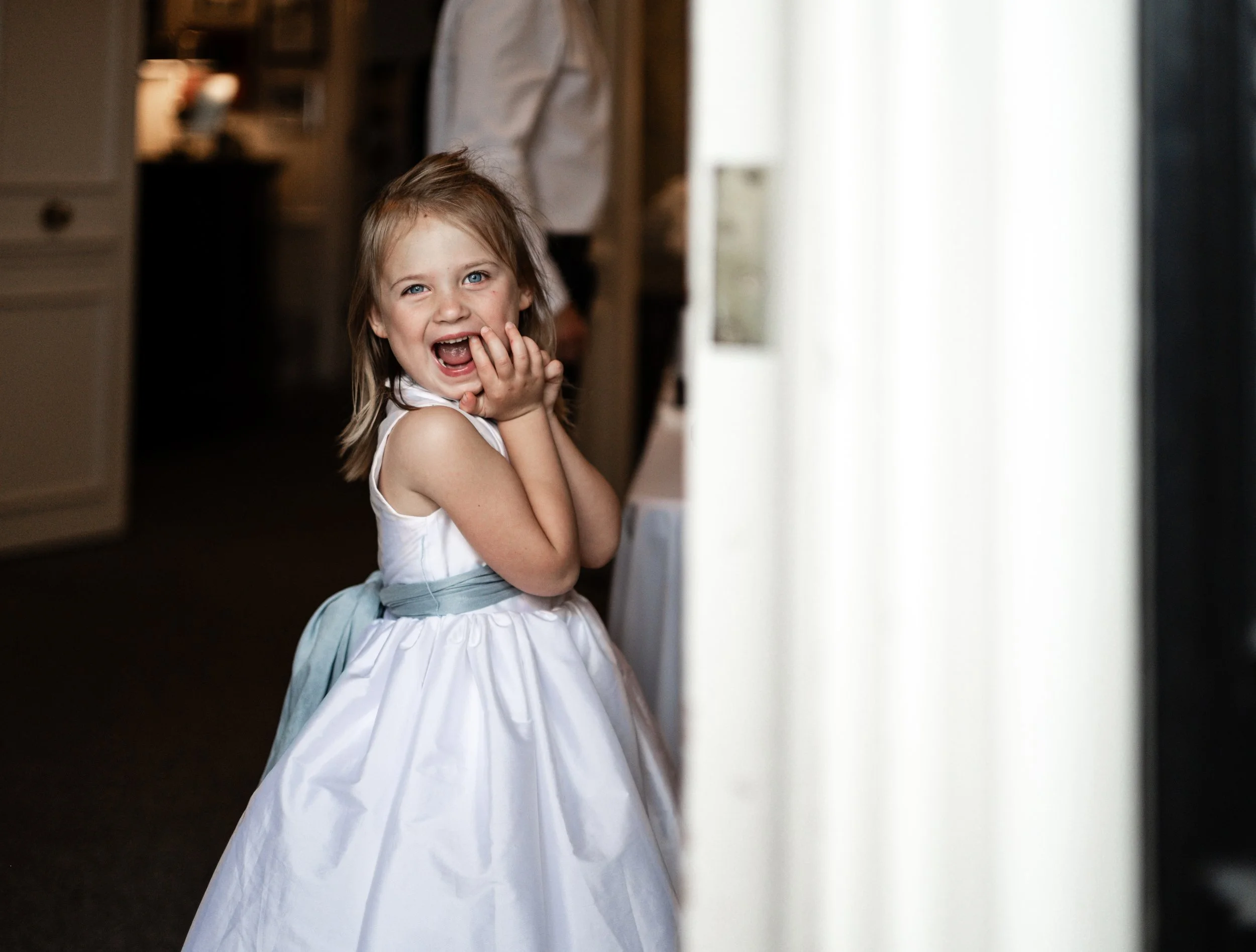 A young girl dressed in a white princess gown, smiling and covering her mouth with her hands, standing near a window with sheer curtains at The Morehead Inn located in Charlotte.