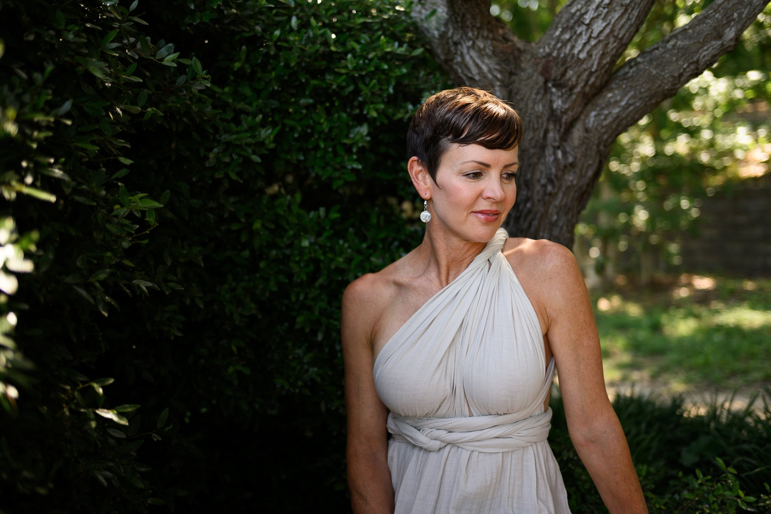 A woman in a white dress with short dark hair and earrings standing outdoors near a tree and green bushes, during actor headshots in Charlotte