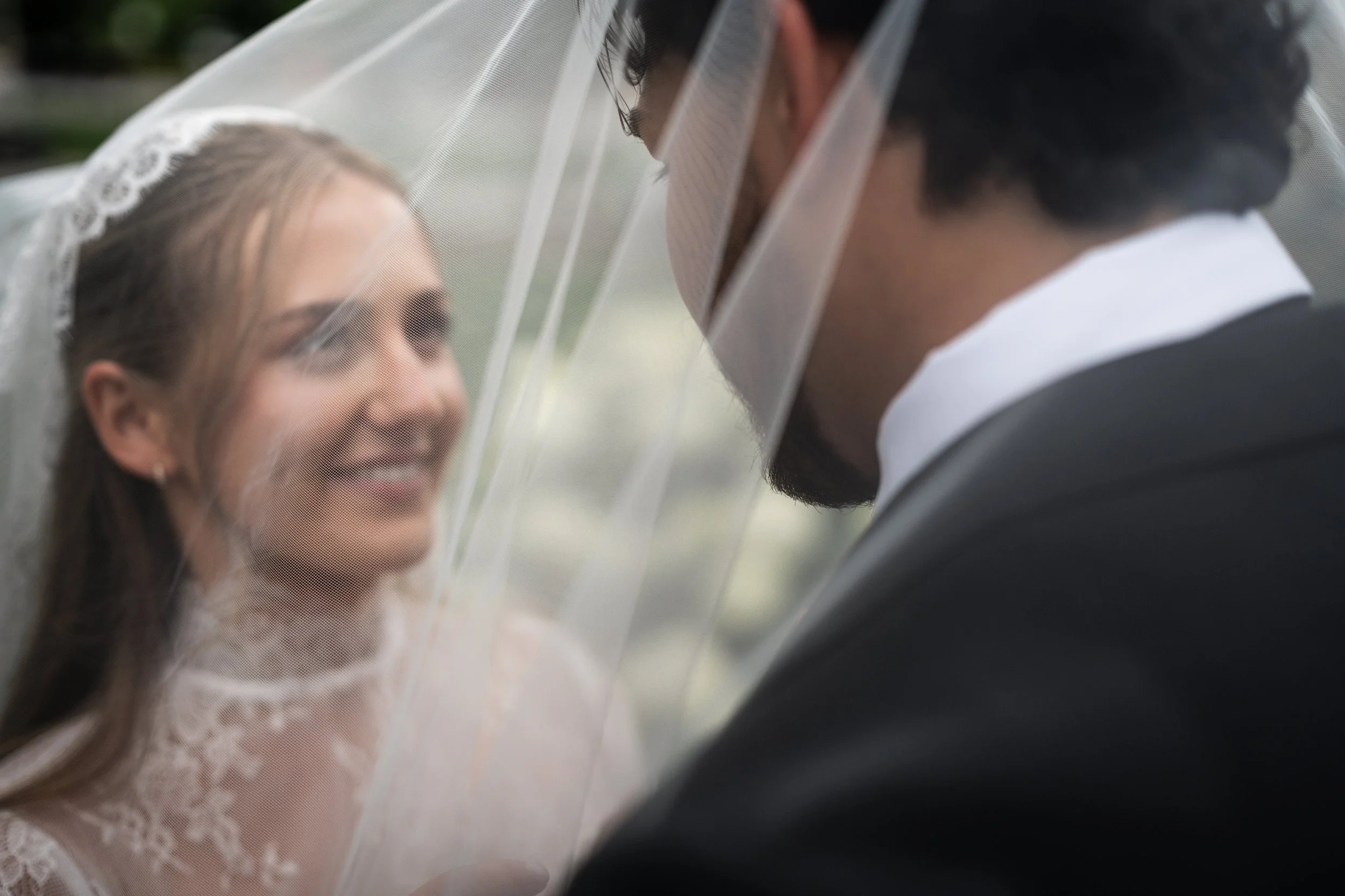 Close-up of a bride and groom face-to-face, separated by a wedding veil, with the bride smiling and the groom leaning in for a kiss at St. Mary's Chapel in Charlotte
