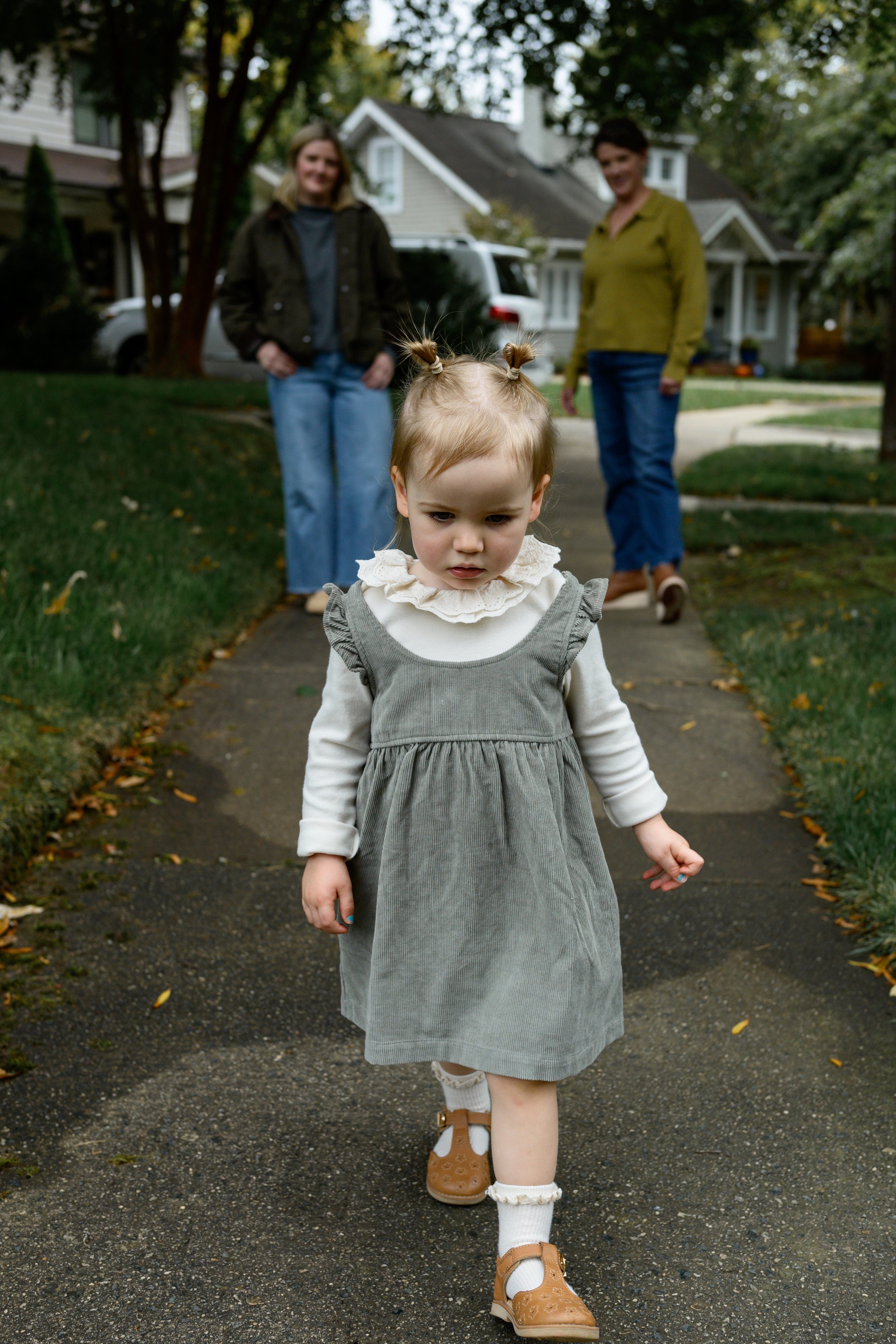 A young girl with blonde hair styled in two buns, walking alone on a sidewalk, with two women standing in the background near houses and trees.