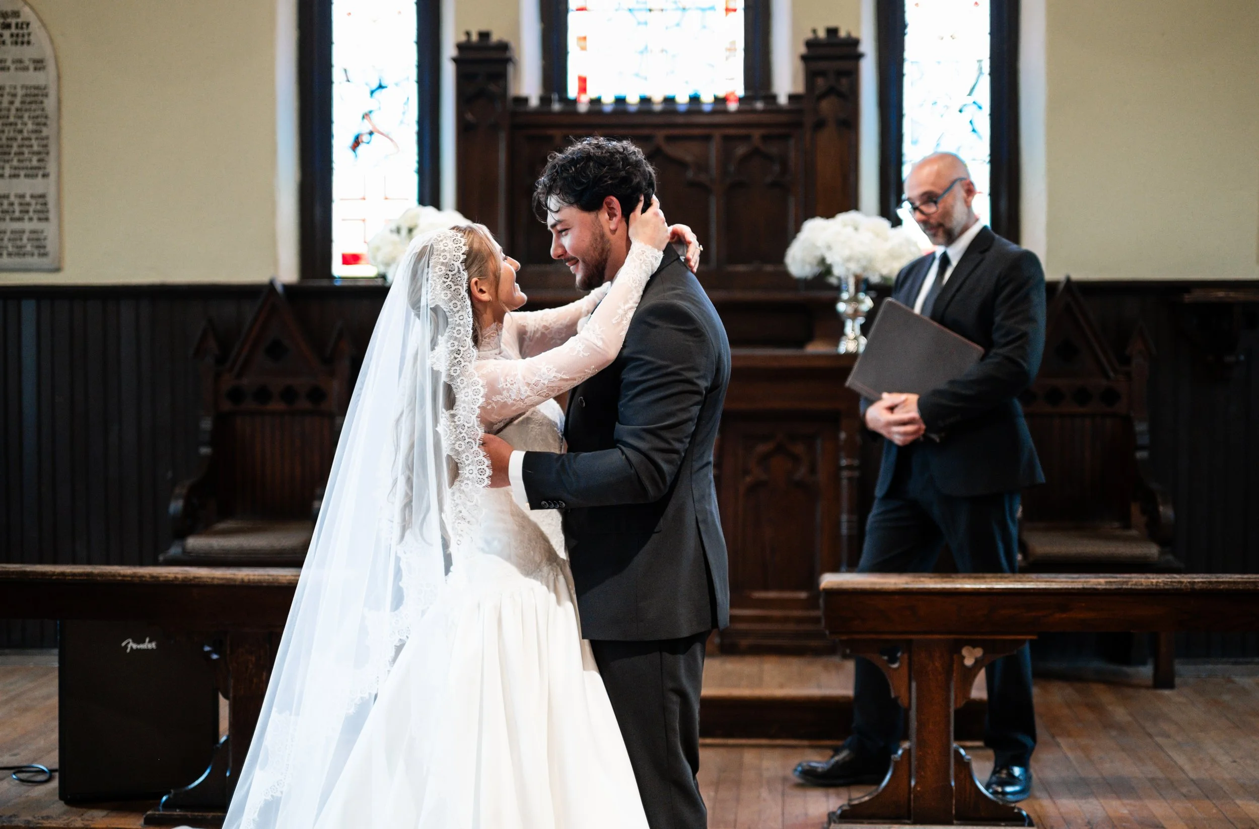 A bride and groom exchange vows and smile at each other during a wedding ceremony inside St. Mary's Chapel in Charlotte, with a male officiant on the right holding a book and smiling.