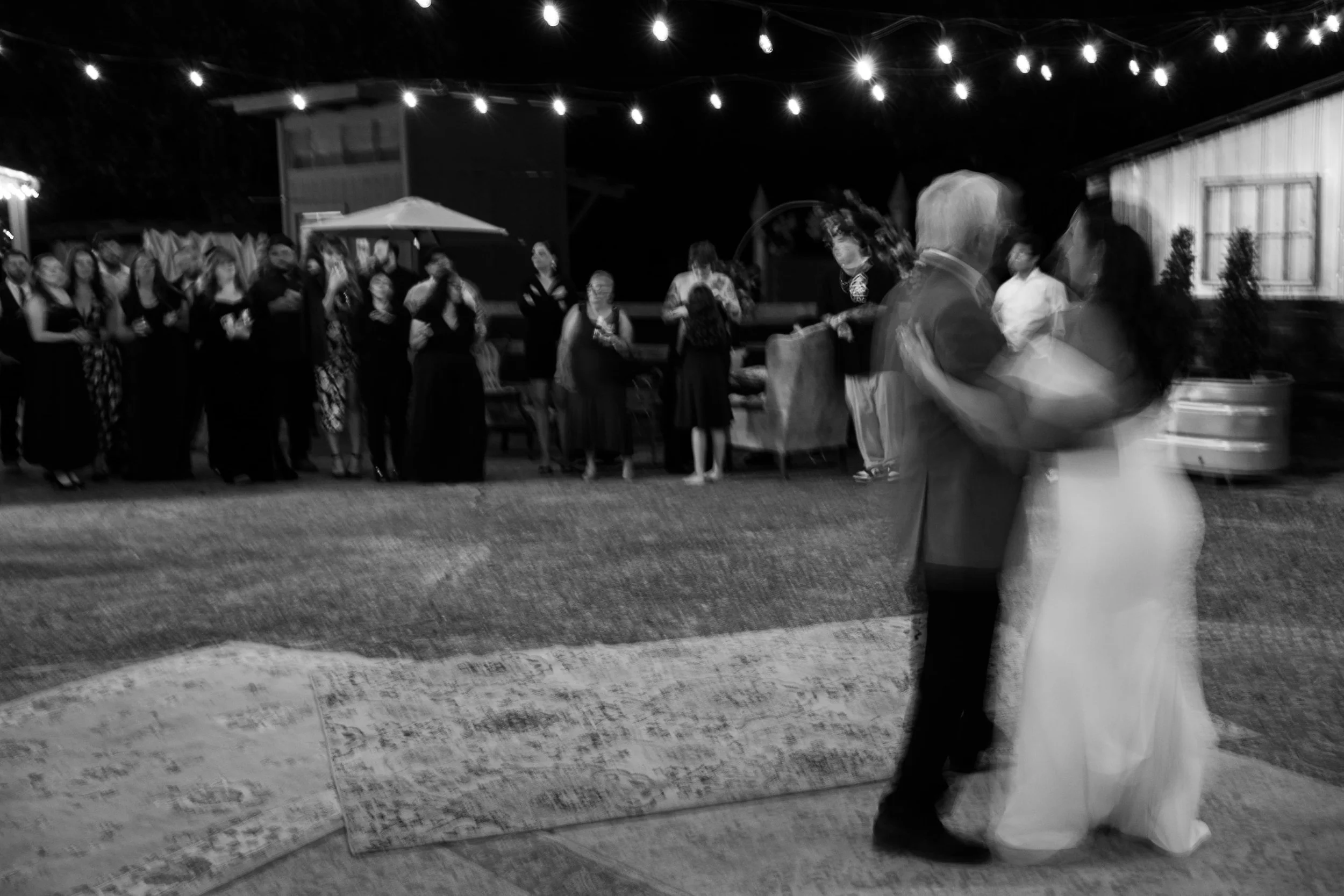 A black and white photo of a wedding reception with a bride and groom dancing in the foreground, blurred, at night under string lights, with guests standing and observing in the background.