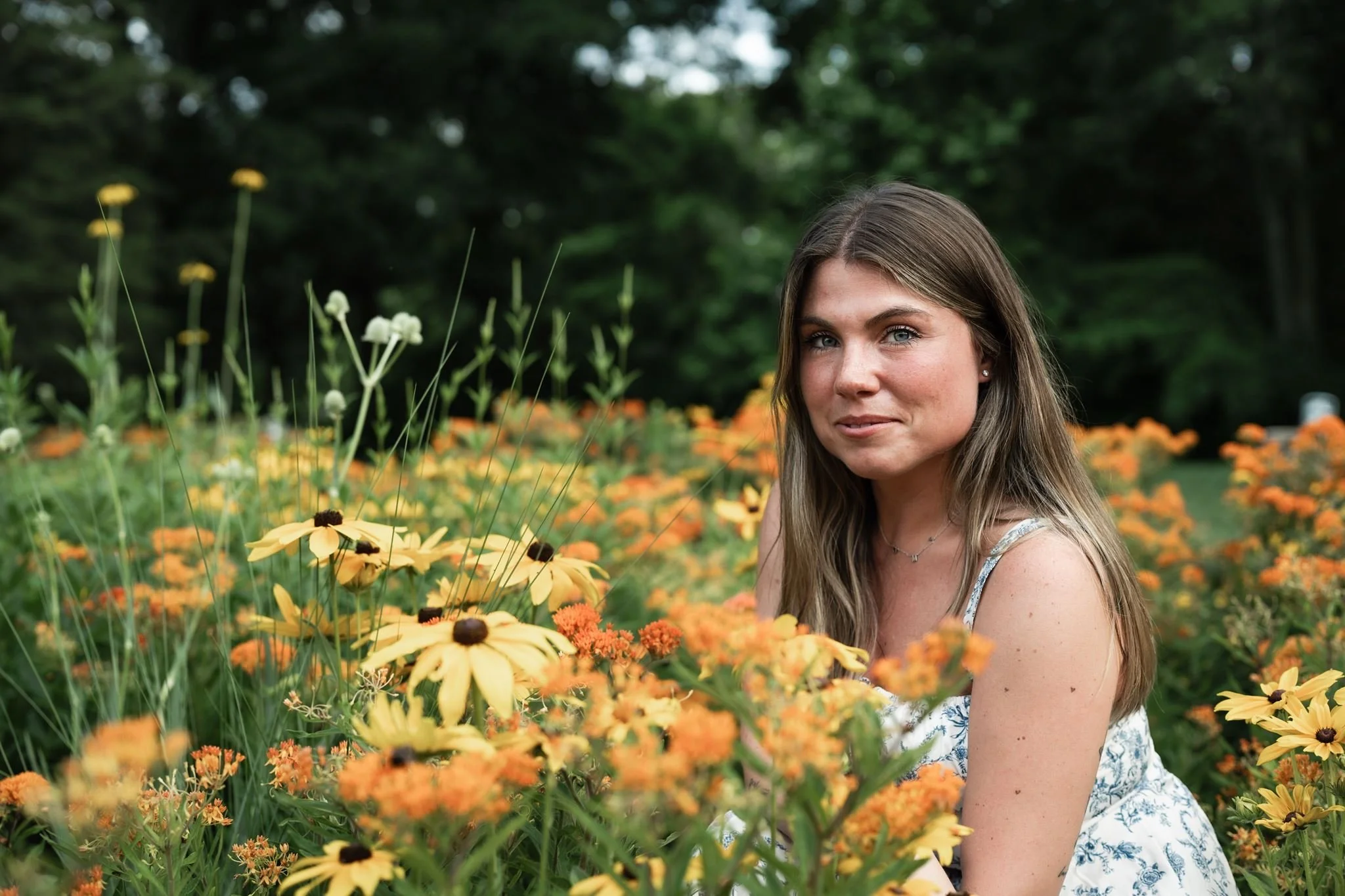 A young woman with long, light brown hair and blue eyes in a field of yellow and orange flowers, with a leafy green background for a portrait photography shoot in Charlotte