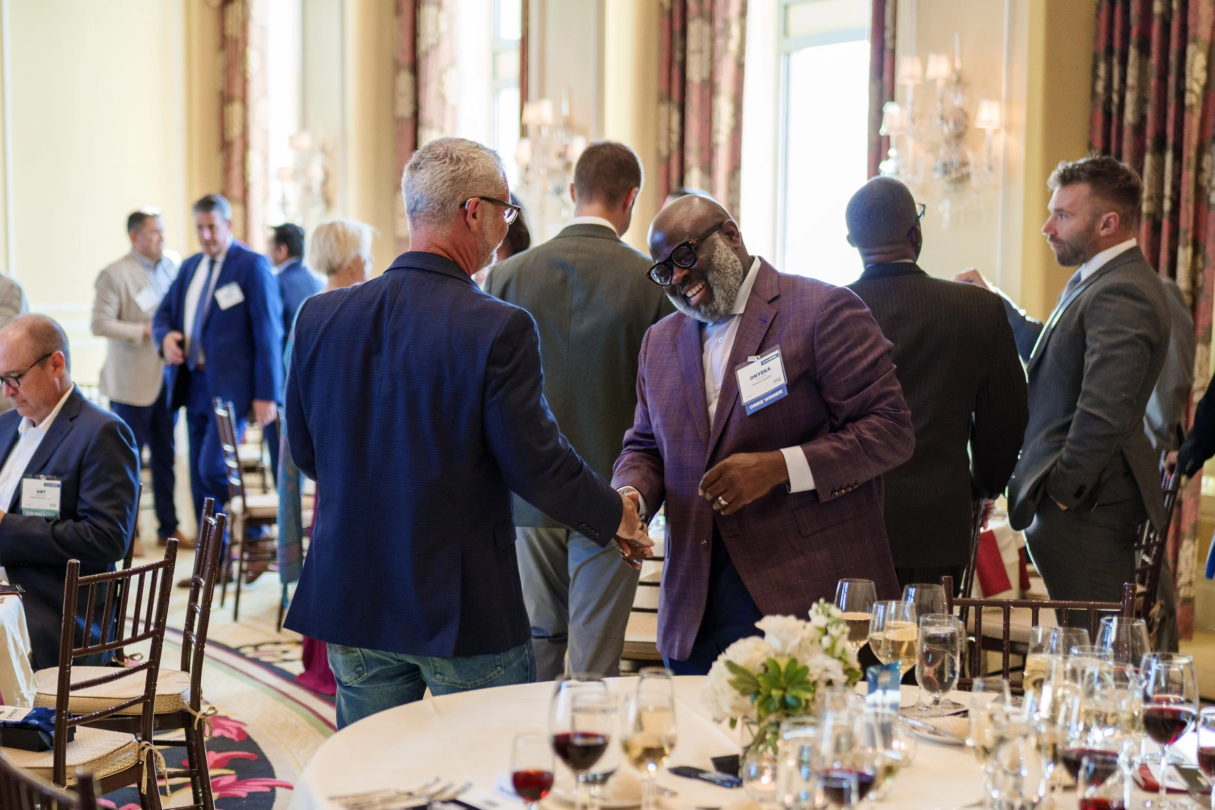 Two men shaking hands and smiling at a professional conference or event. One man wears a blue checkered blazer and glasses, and the other wears a purple blazer and glasses. In the background, other attendees are mingling in a well-lit, decorated room