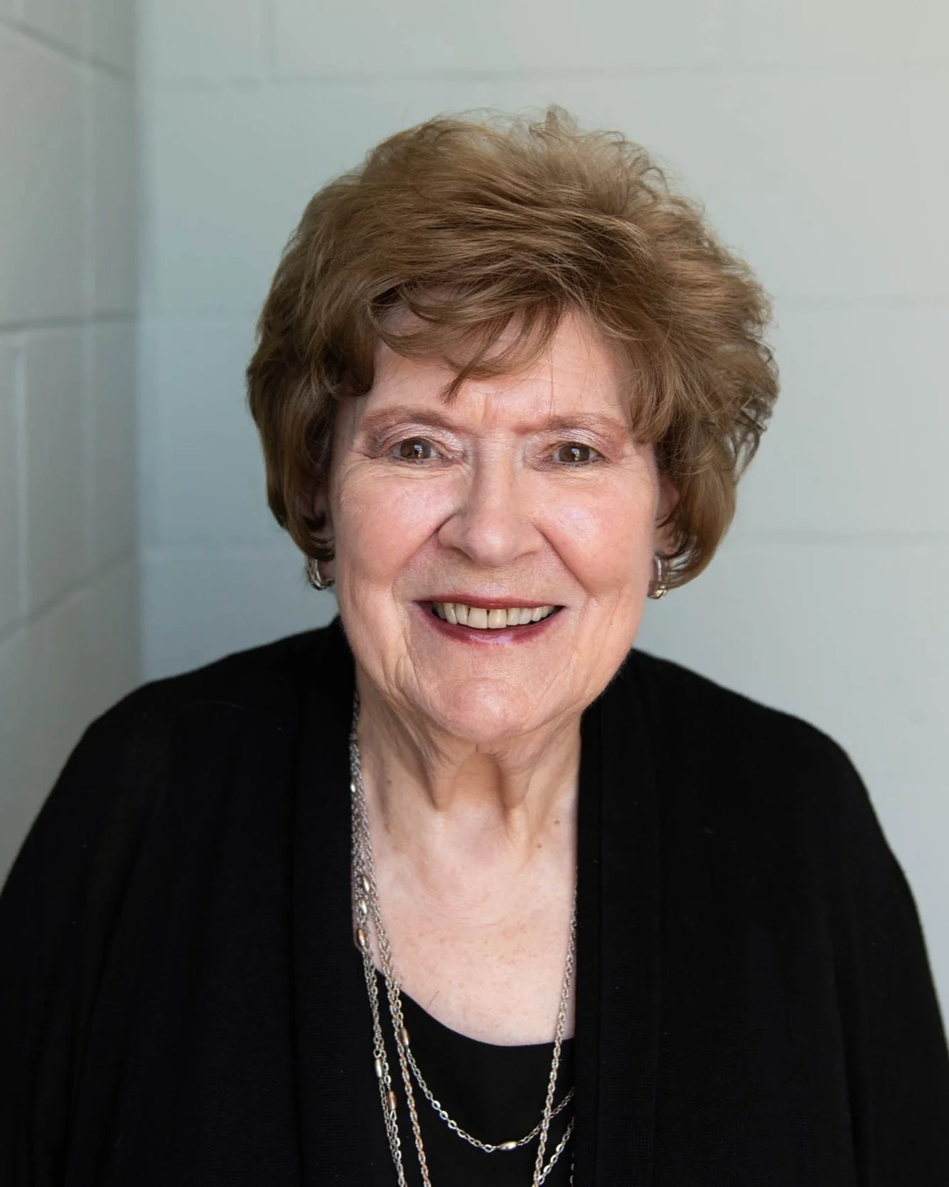 A smiling elderly woman with short, curly gray hair, wearing a black top and layered silver necklaces, in front of a light-colored wall.