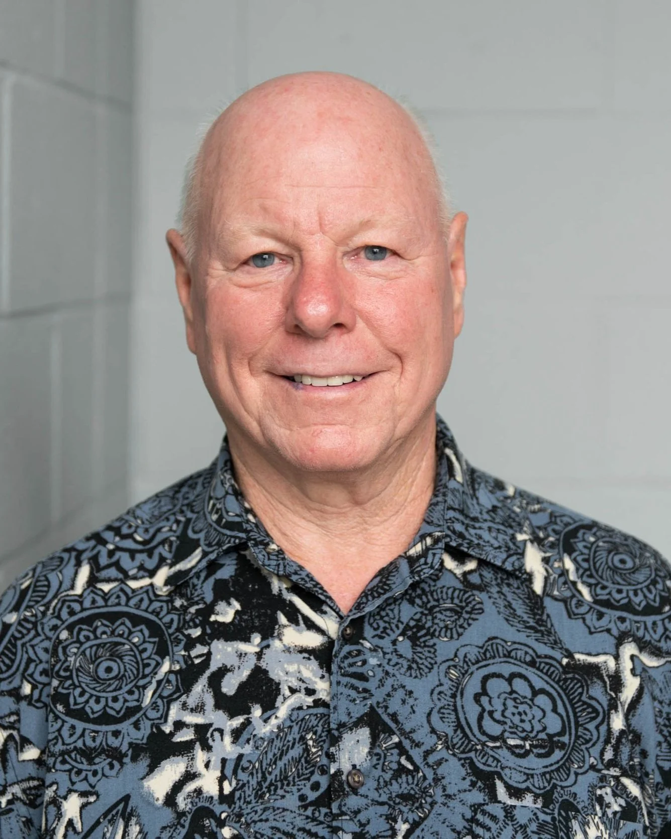 A smiling older man with a bald head and blue eyes, wearing a patterned button-up shirt, standing against a light gray background.