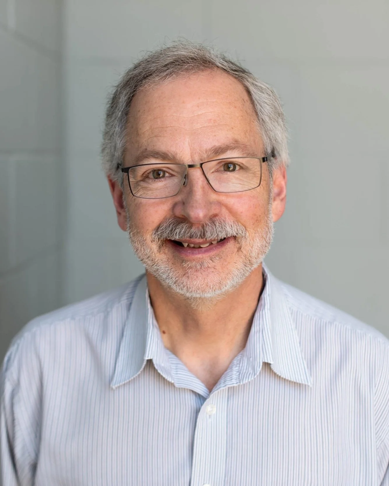 A smiling older man with gray hair and a beard, wearing glasses and a light blue striped shirt, standing in front of a neutral background.