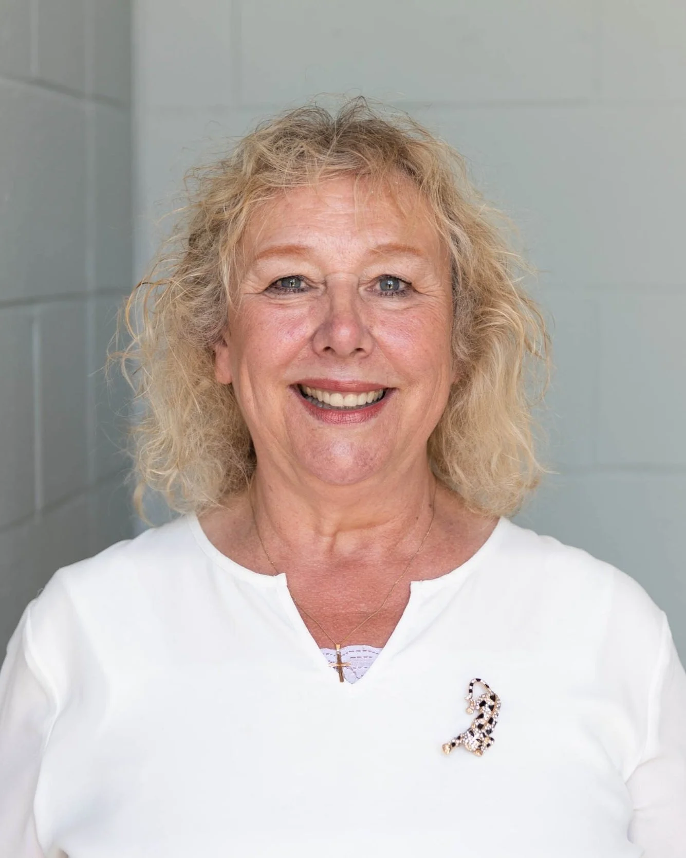 A smiling middle-aged woman with curly blonde hair wearing a white top with a leopard print brooch and a small gold cross necklace, standing in front of a pale wall.