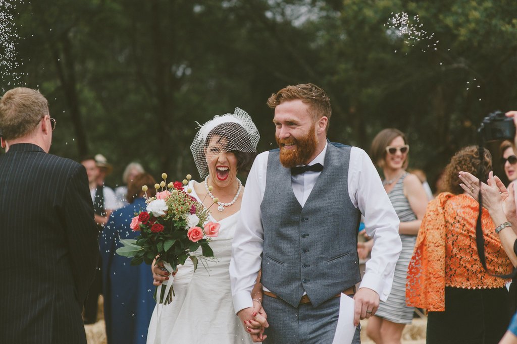A bride and groom smiling and holding hands outdoors during their wedding celebration, surrounded by guests.