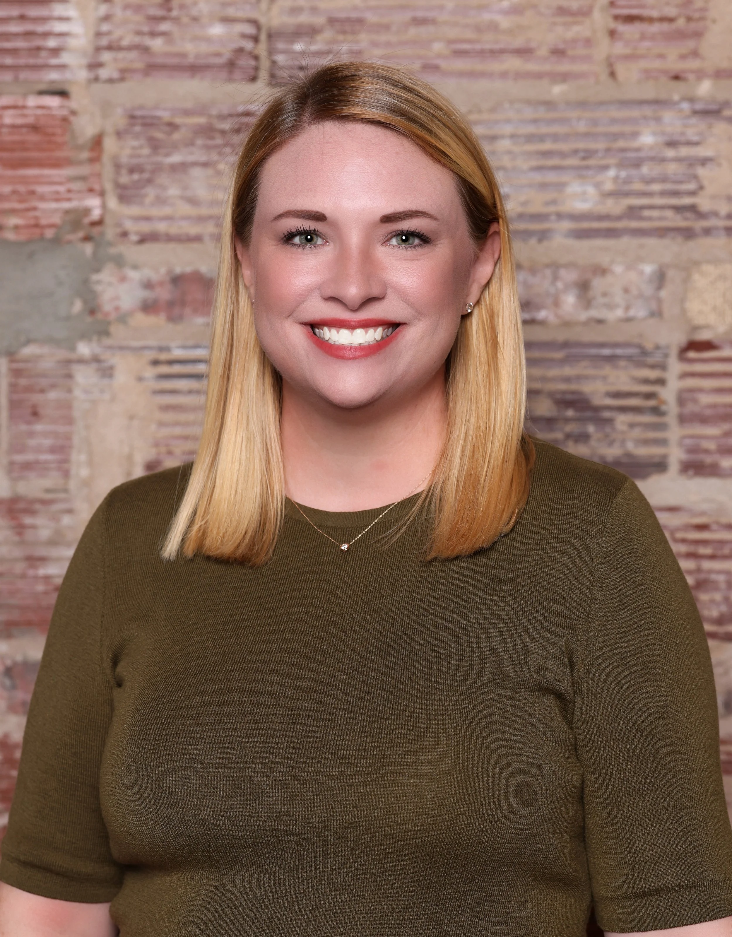 Close-up of a woman with blonde hair, blue eyes, and a bright smile, standing against a textured brick wall background, wearing a dark green top and a delicate necklace.