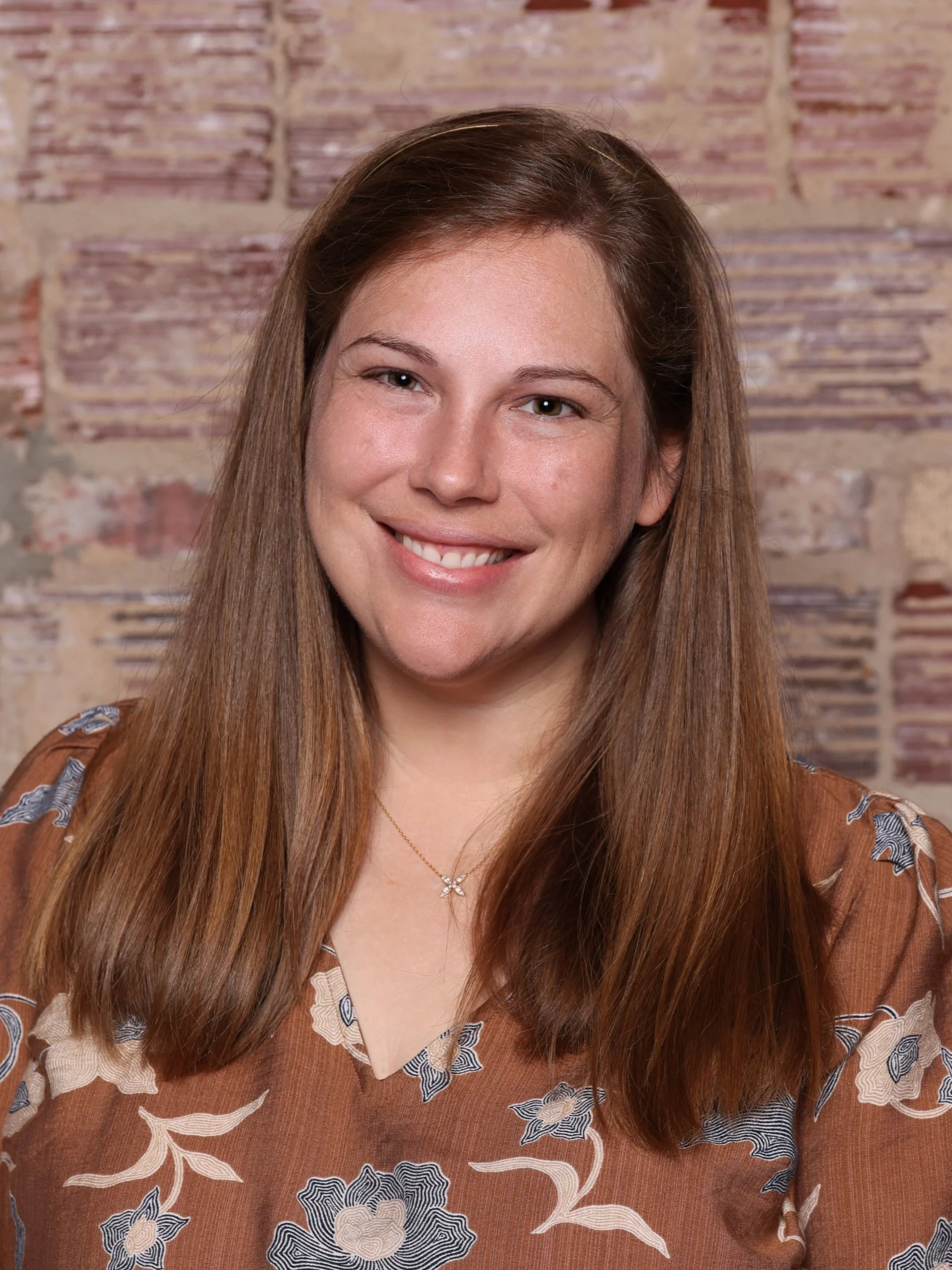 A woman with long brown hair, smiling with white teeth, wearing a black dress with red, gold, and green floral pattern, and a silver headband, standing in front of an exposed brick wall.