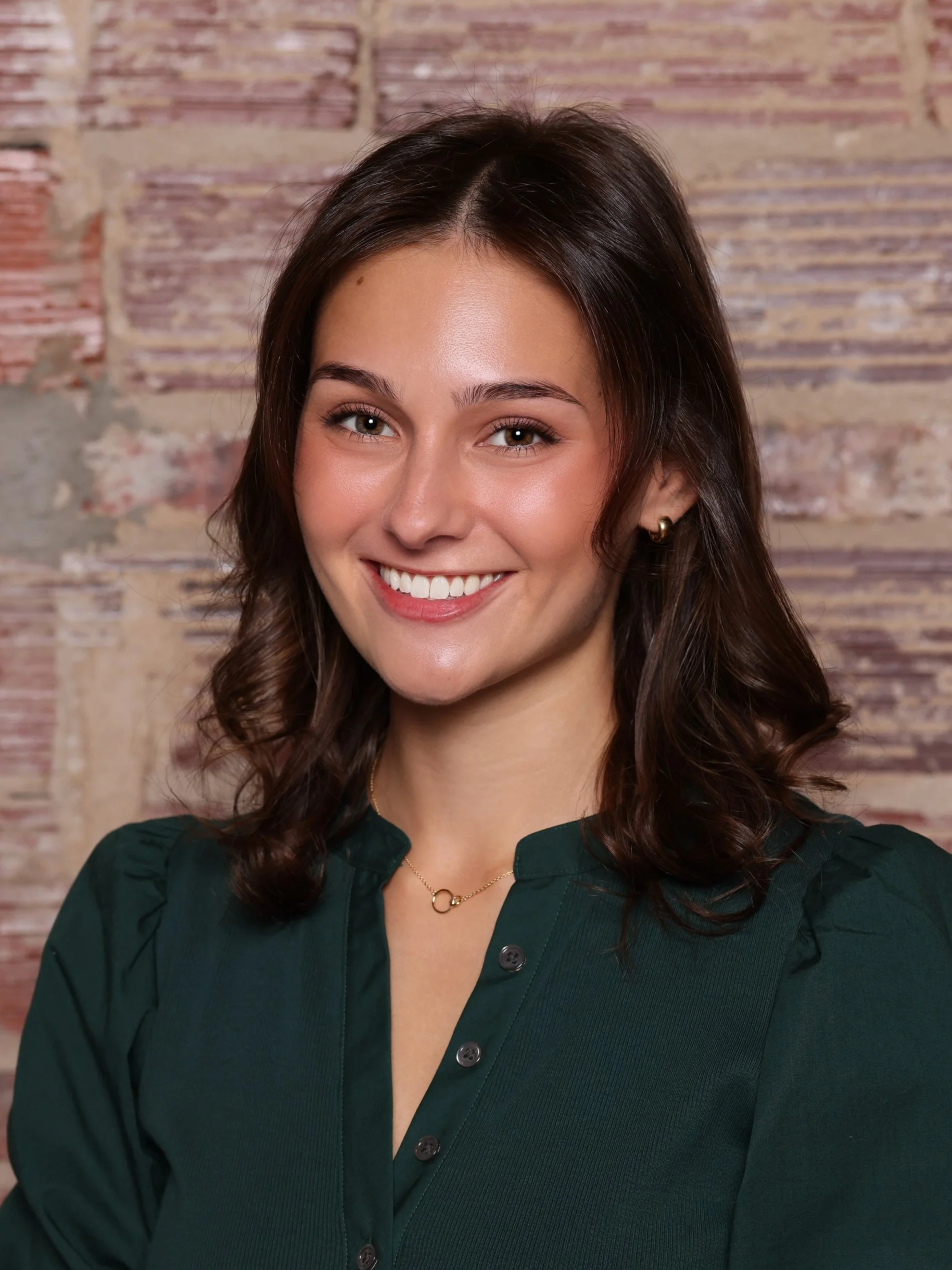 A young woman with shoulder-length brown hair, wearing a floral-patterned sweater, smiling with arms crossed in front of a rustic brick wall background.