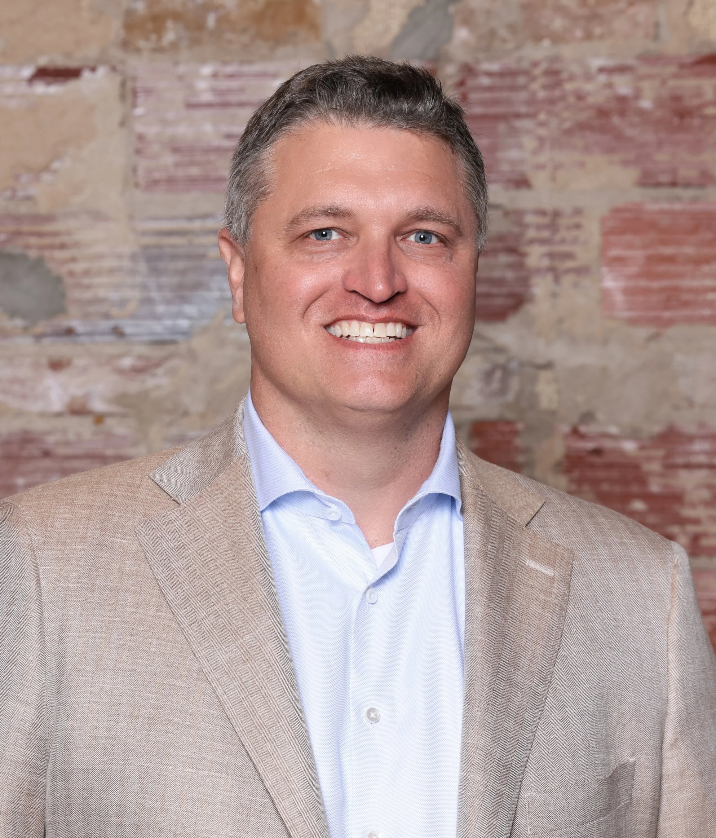 A portrait of a smiling man in a beige suit jacket and light blue shirt standing in front of a brick wall.